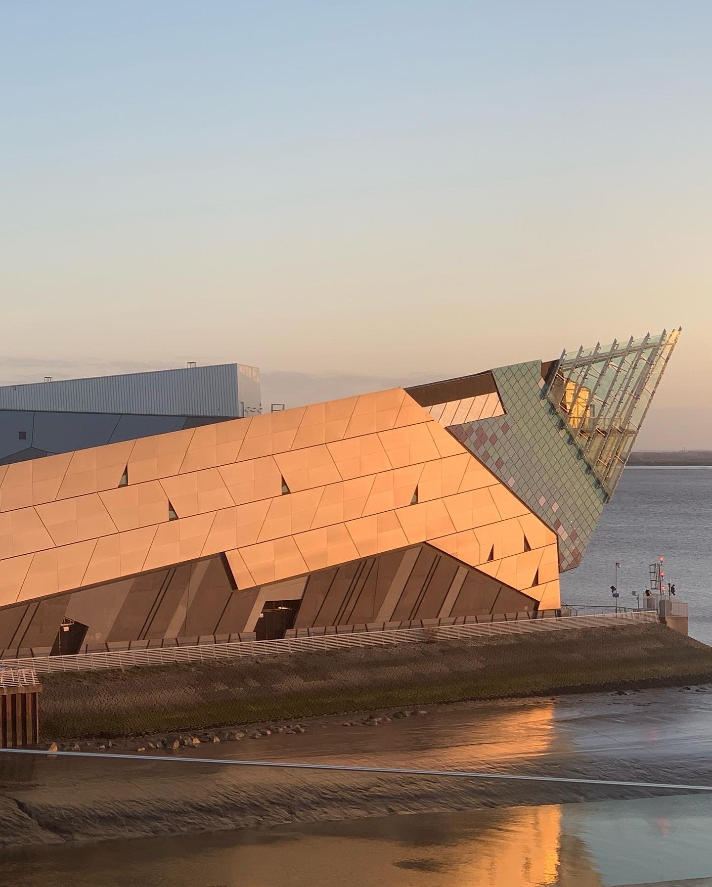The Deep. A modern architectural building with a geometric design, copper-colored panels, and glass sections, near a body of water during sunset.