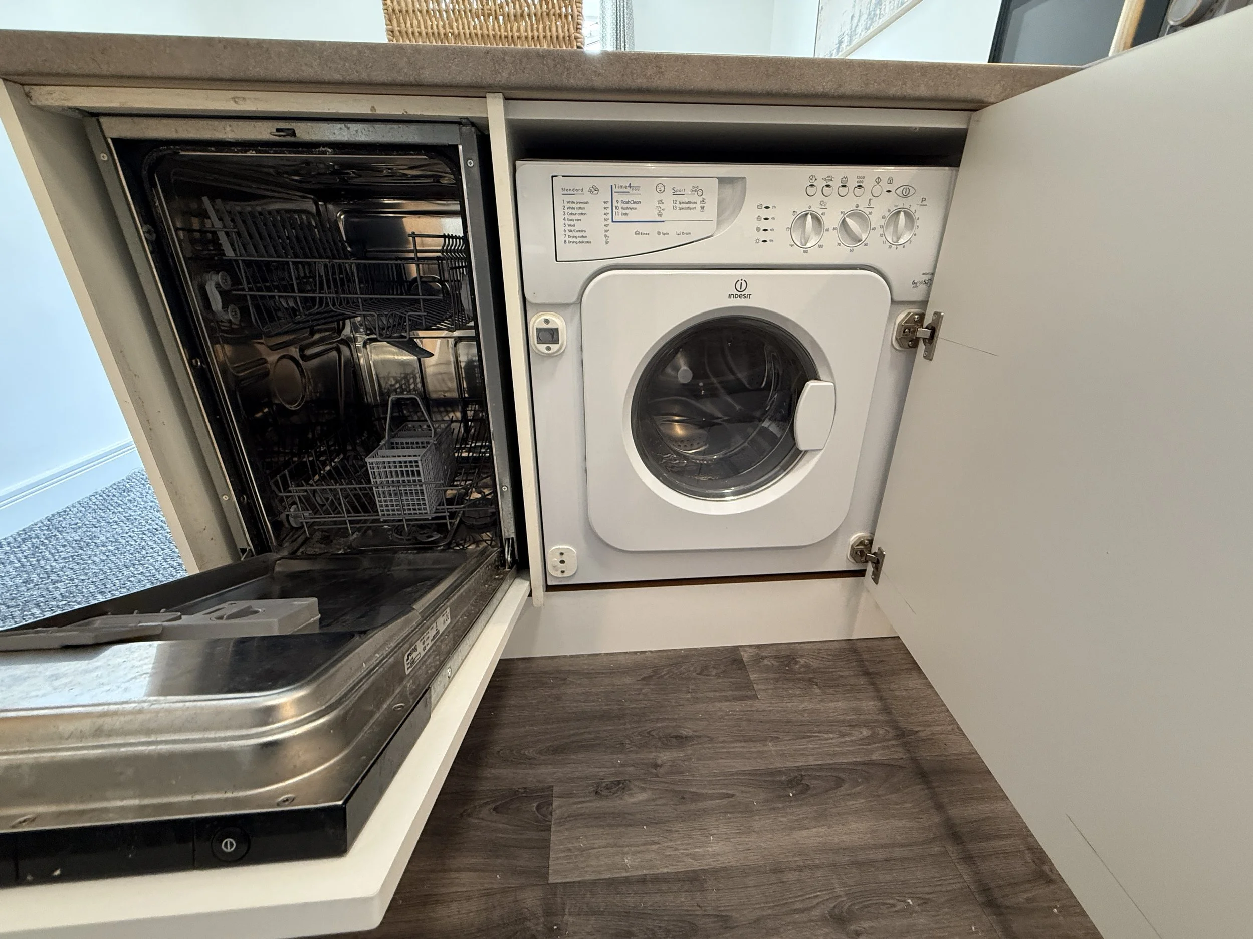 Open kitchen cabinet with dishwasher and washer-dryer, dark vinyl flooring, and countertop.