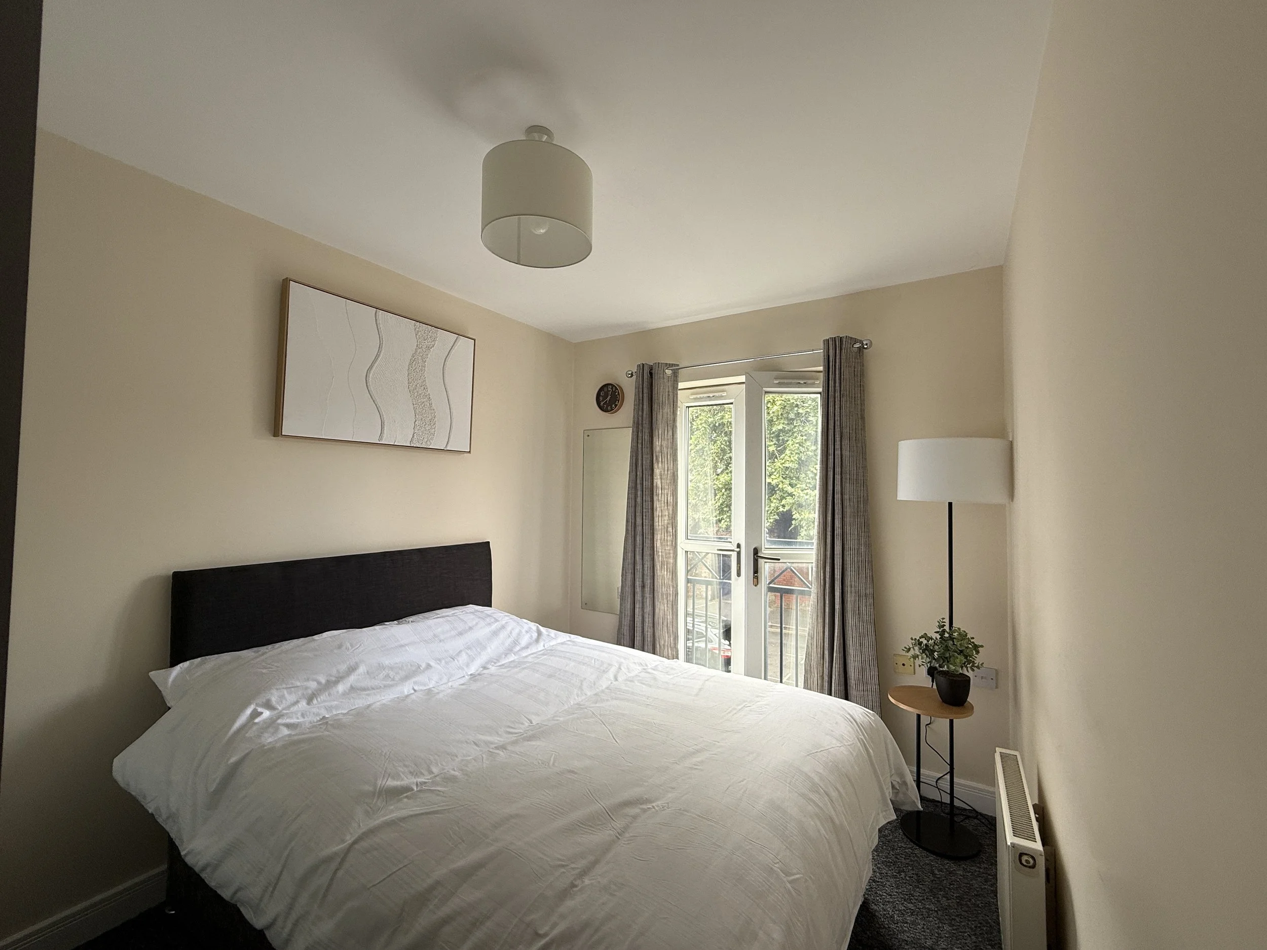 A bedroom with a bed covered in white bed sheets, a nightstand with a small plant, a floor lamp, a window with curtains, a wall clock, and framed artwork on the wall.