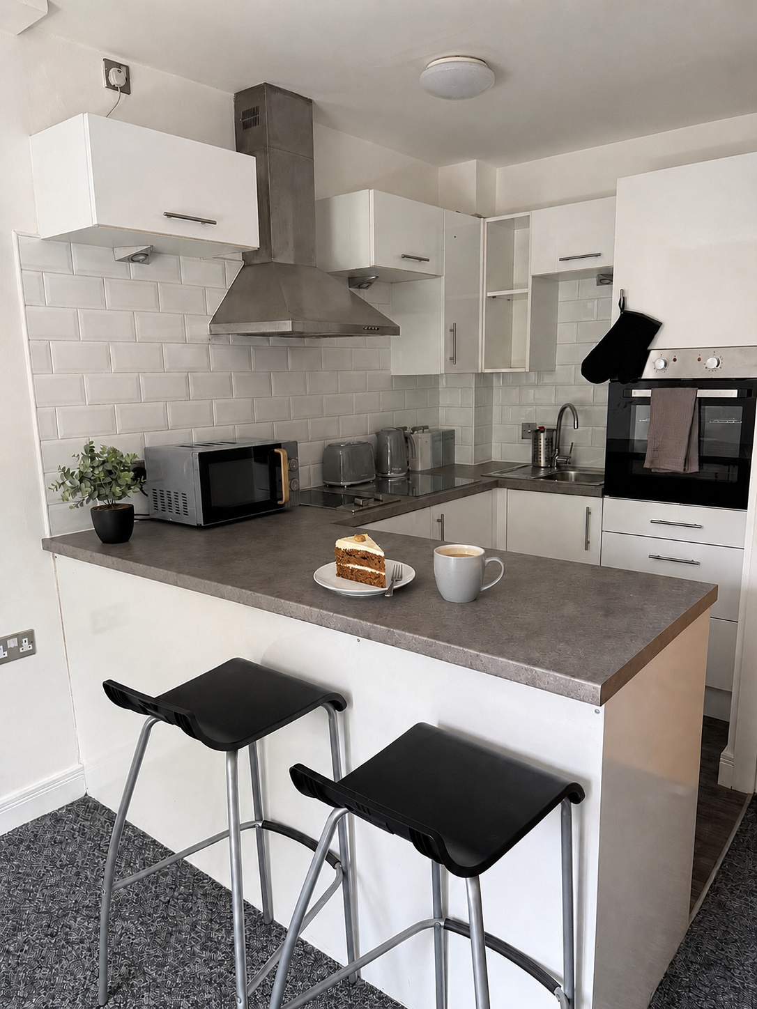 Modern kitchen with white cabinets, stainless steel range hood, microwave, toaster, and kettle on grey countertop. There are two black bar stools at the counter, with a slice of cake and a coffee mug on it.