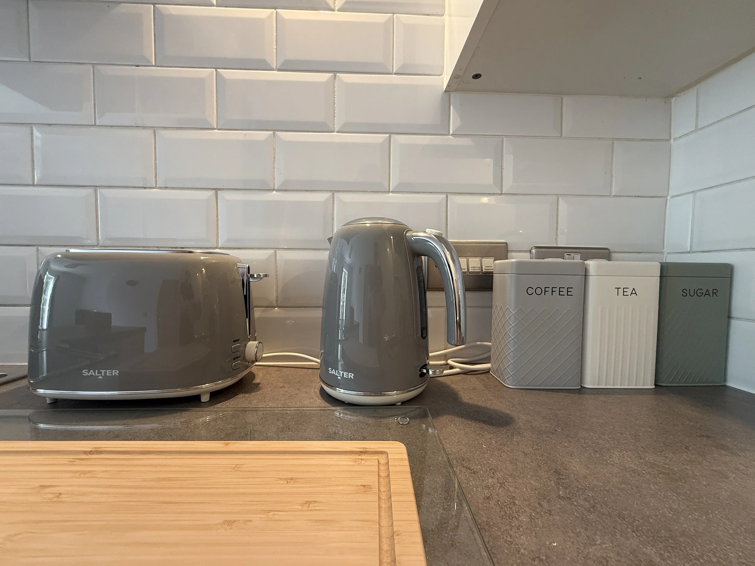 Kitchen countertop with a grey toaster, kettle and three white coffee canisters labeled coffee, tea, and sugar. Wooden cutting board in the foreground and white subway tile backsplash.