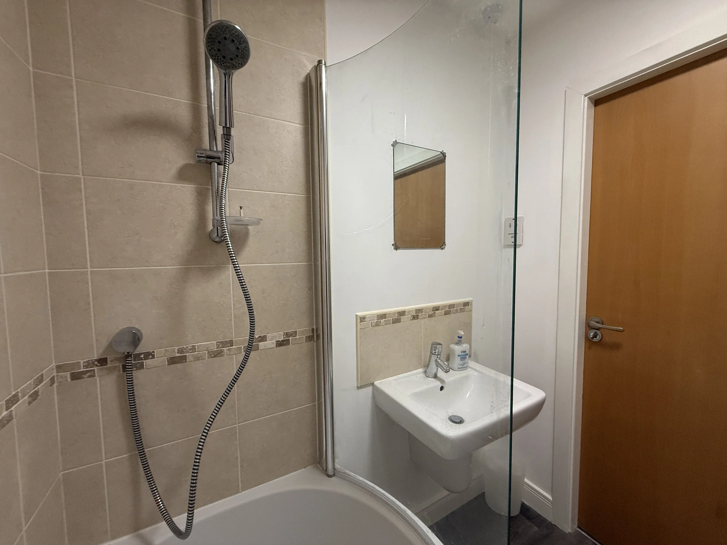 Bathroom with a corner shower, beige tiled walls, a small mirror above a white sink, and a door.