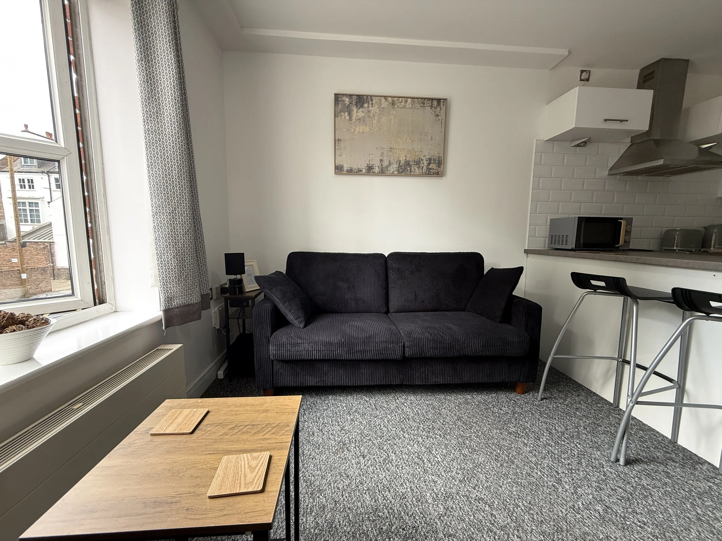 A living room with a large window, a grey/black sofa, a wall painting, a small side table with a lamp, a kitchen area with white cabinets, a microwave, and two bar stools.
