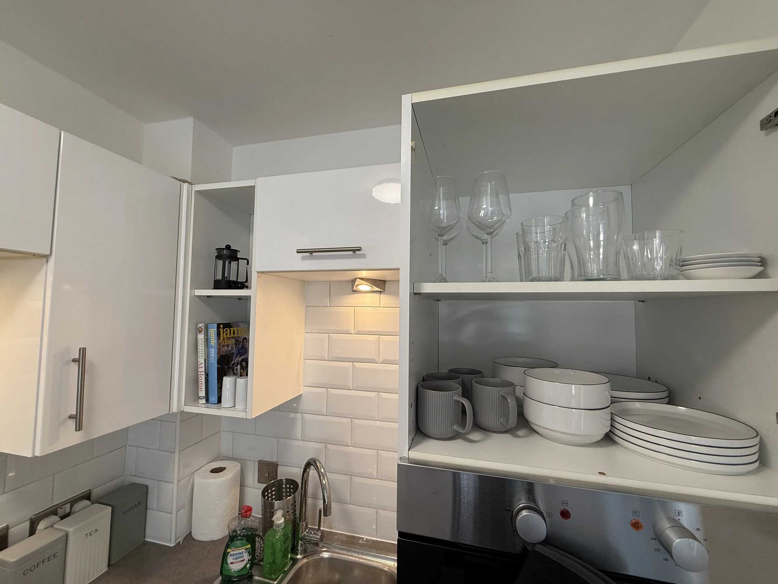 A kitchen cabinet with various glasses and plates, alongside cleaning supplies near the sink on the right.
