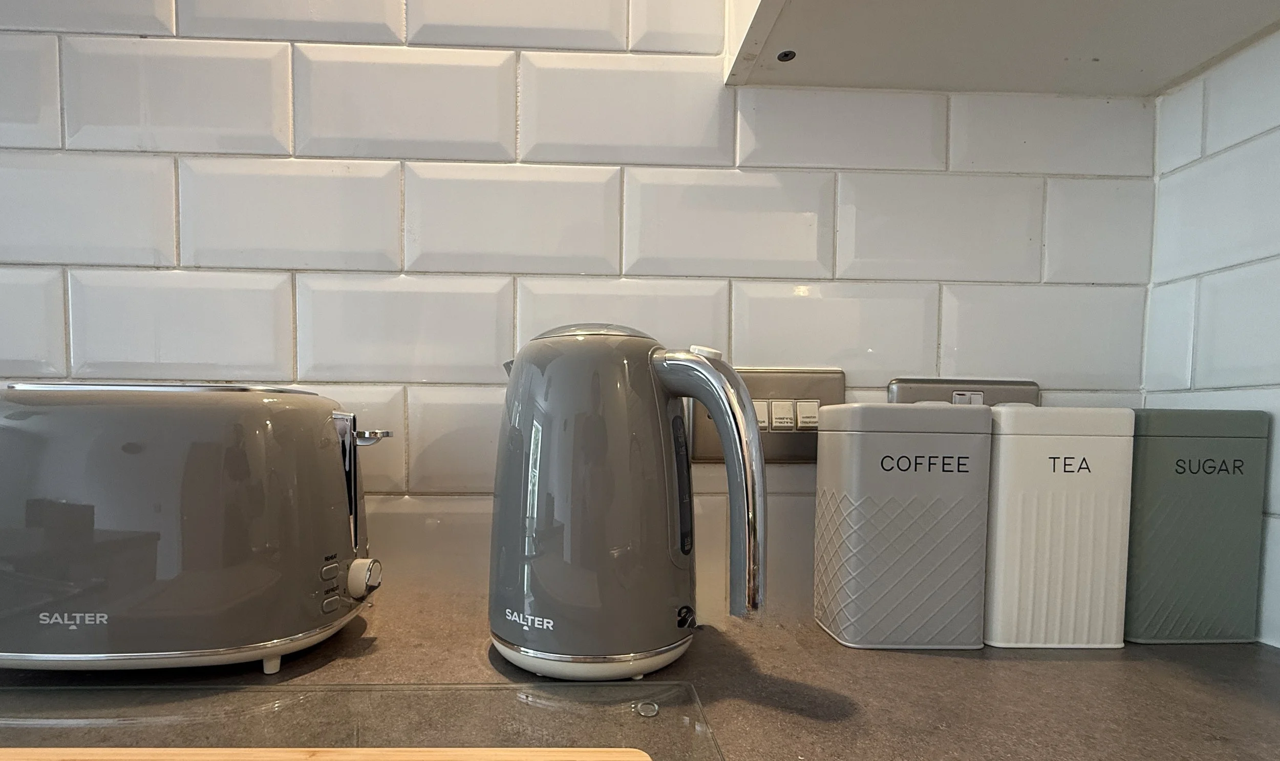 A kitchen countertop with two grey electric kettle toasters, and three labelled containers for coffee, tea, and sugar against a white tiled backsplash.