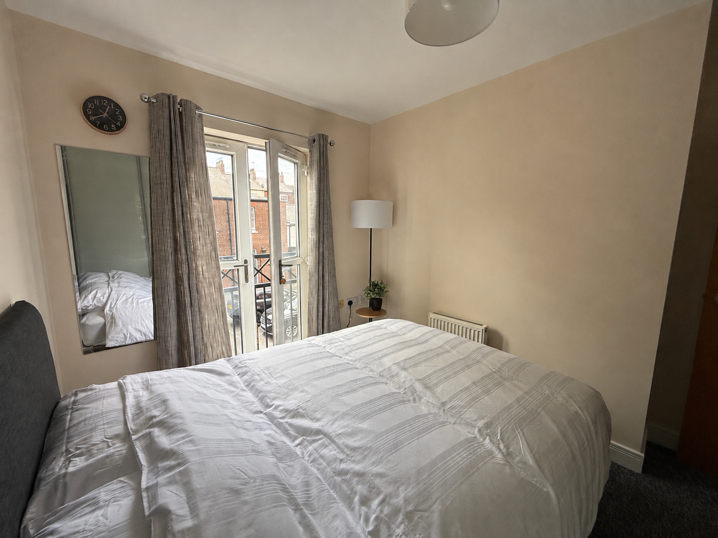 A bedroom with a bed covered in a white striped duvet, a full-length mirror on the wall, a window with beige curtains, a radiator, a small side table with a potted plant, and a white floor lamp.