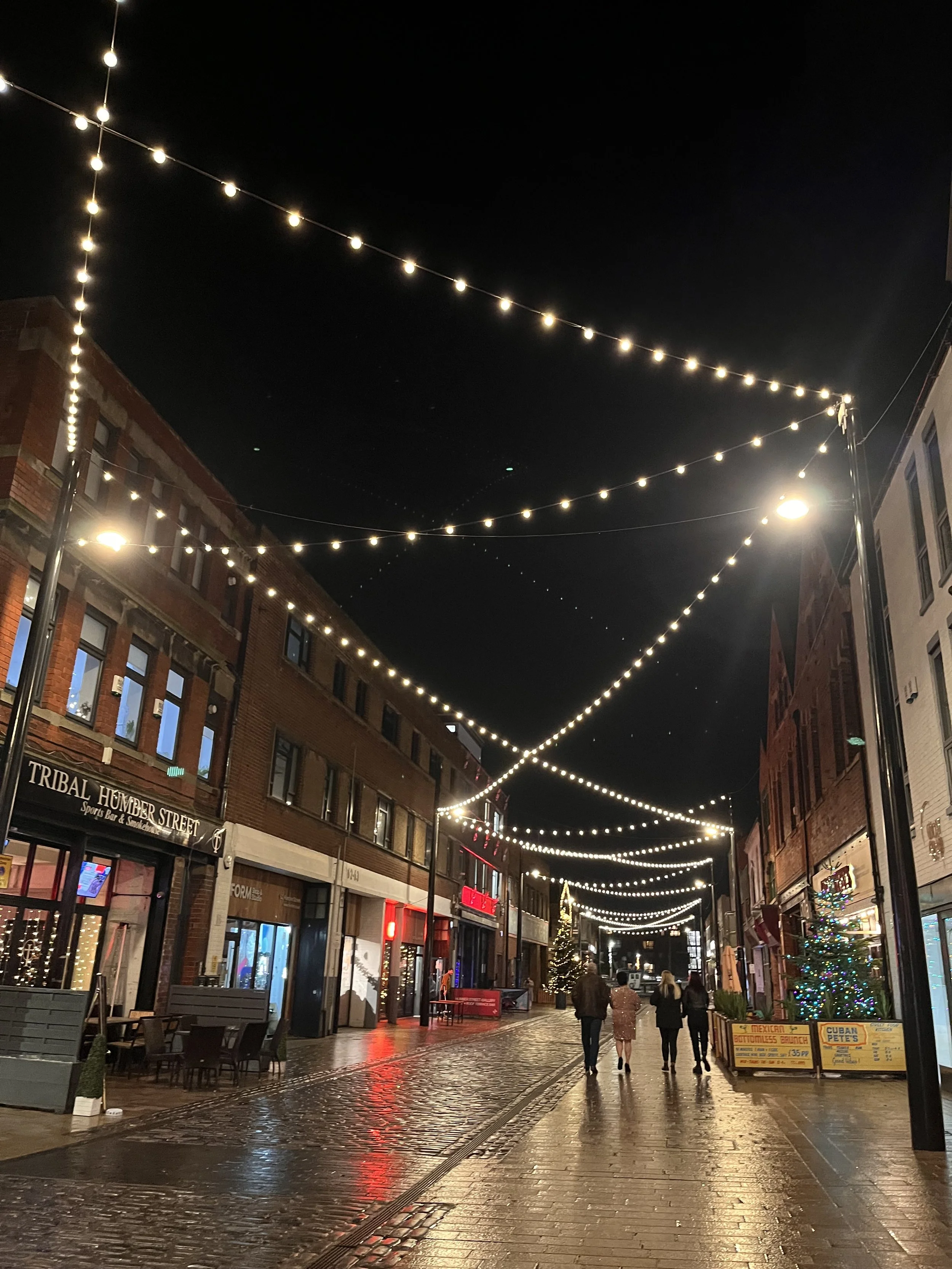 A nighttime street scene of Humber Street, decorated with string lights overhead, with people walking and shop signs visible, including a Christmas tree with lights.