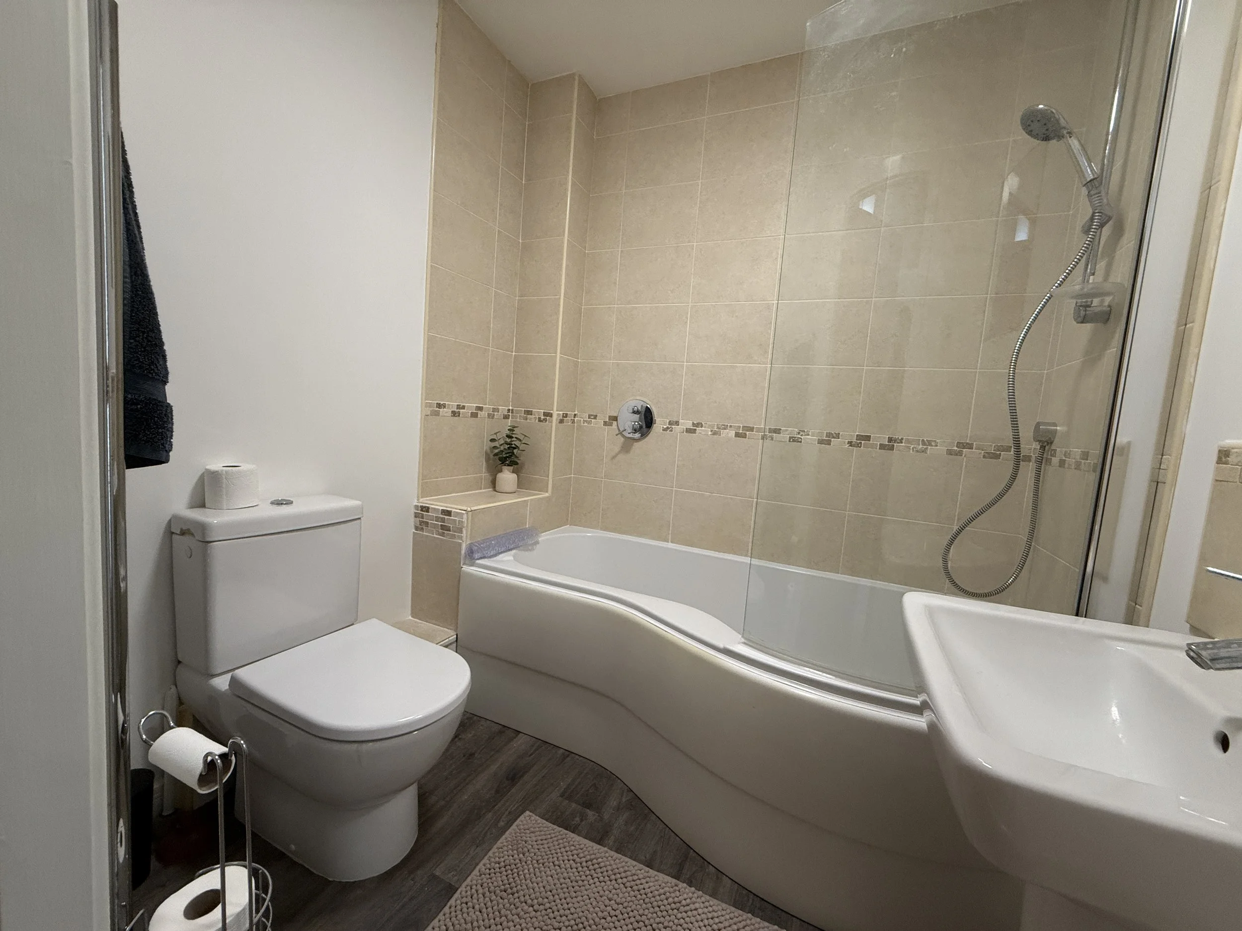 Bathroom with a toilet, bathtub, beige tiled walls, and a shower. There is a small plant on a tiled ledge and a toilet roll holder.