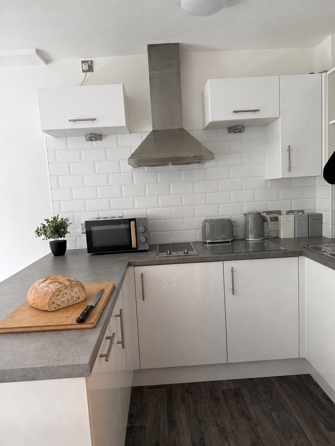 Modern kitchen with white cabinets, grey countertops, a stainless steel range hood, toaster, kettle, microwave, and jar containers, with a loaf of bread on a cutting board.