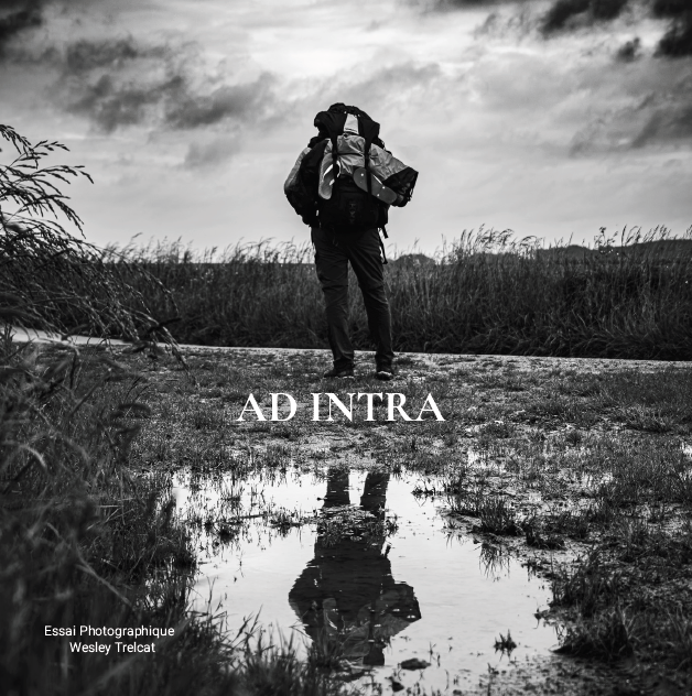 Une personne portant un sac à dos marche dans une zone de marais ou de terrain humide, avec des nuages dans le ciel. La photo est en noir et blanc et le reflet de la personne est visible dans l'eau.