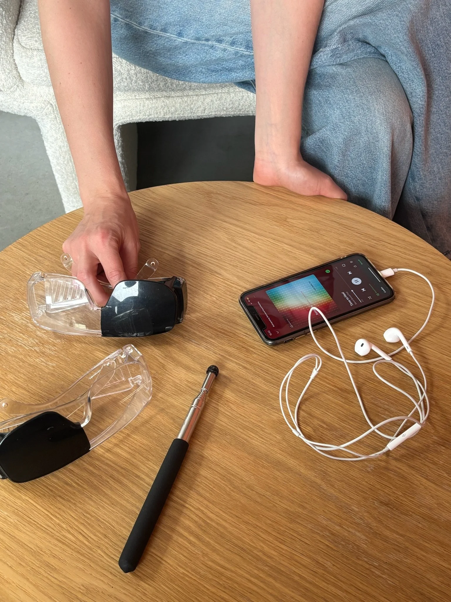 Een houten tafel met verschillende elektronische apparaten en accessoires, waaronder een smartphone met koptelefoon, een soldeerbout, een zwarte tracker, en een borstel. Een persoon met een blauwe broek en een lichtgrijze trui zit aan de tafel.