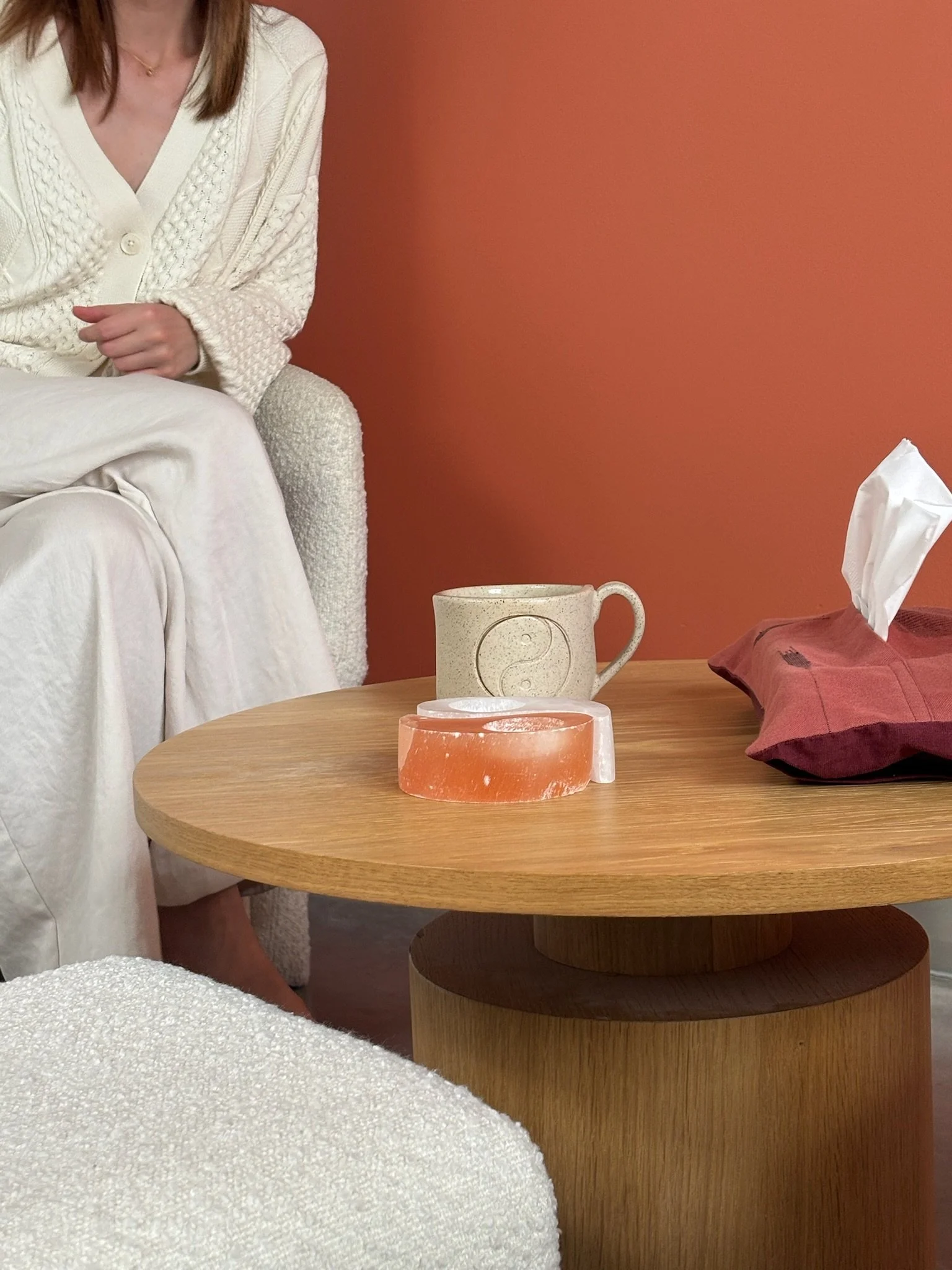 Een houten salontafel met een keramieke mok met yin-yang symbool en een kopje zeep. Op de tafel ligt een geruïneerde donsdeken. Op de achtergrond een rood/oranje muur en een nagellakdoekje.