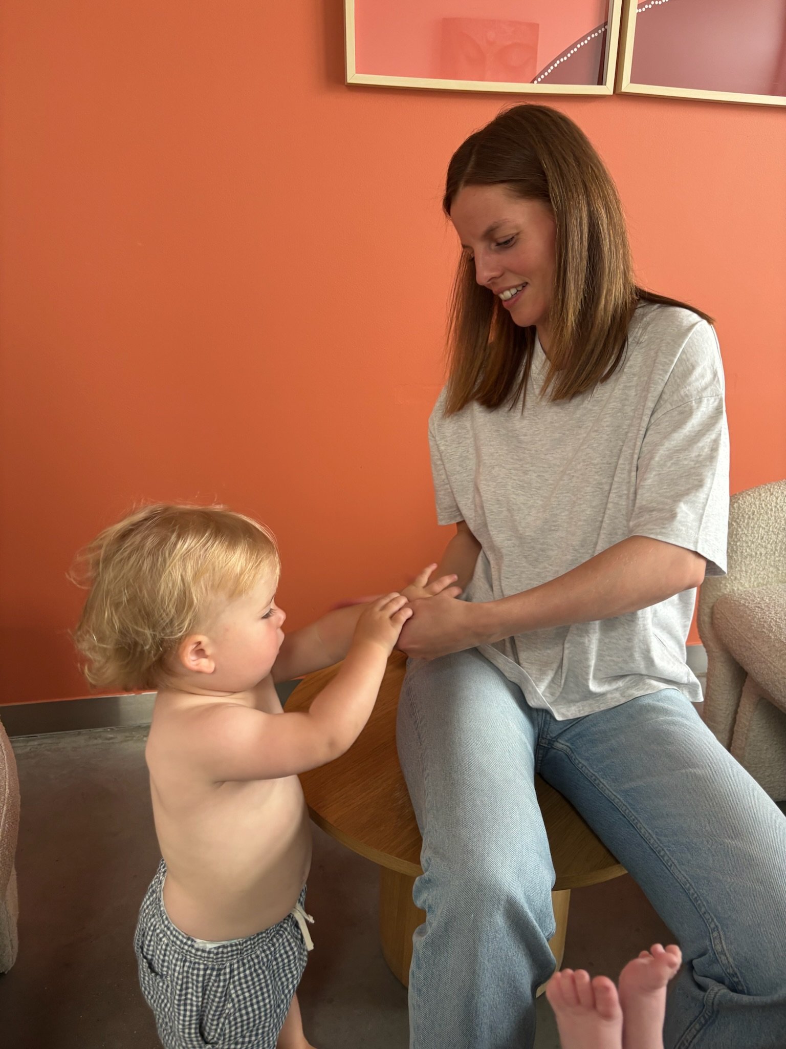 Een jonge jongen met blond haar, die een hand vasthoudt van een vrouw met bruin haar, die glimlacht en op een stoel zit. De achtergrond is een oranje muur met twee framed kunstwerken.