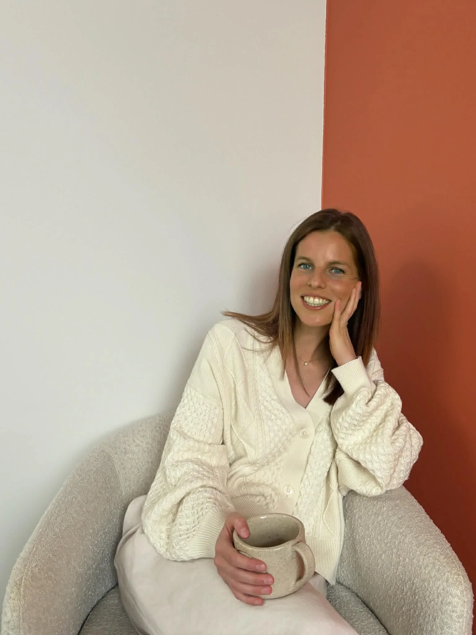Vrouw zit in een beige stoel, houdt een grote keramieken mok en glimlacht naar de camera. Ze draagt een witte kabeltrui en zit tegen een muur met een drie-kleurenschema: wit, beige en terracotta.