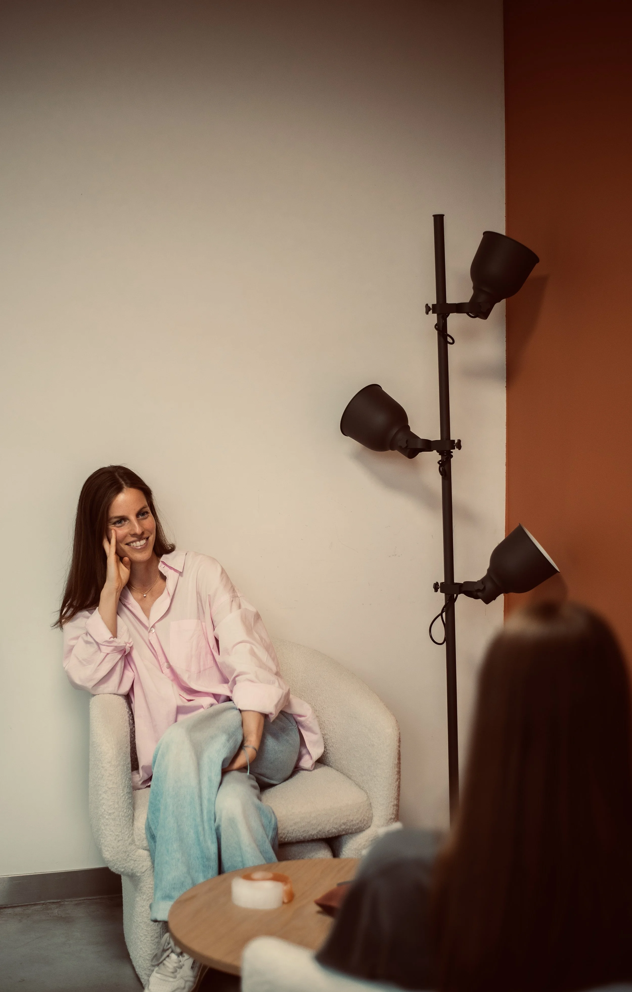 Vrouw zit op een comfortabele witte bank, lachend en met haar hand op haar gezicht, terwijl ze in een gesprek is. Ze draagt een roze blouse en lichte spijkerbroek, met een deken over haar benen. In de achtergrond hangt een muur met een statische kleur, en er staat een lamp met drie matzwarte spotlights gericht naar haar toe. Een persoon voor haar, waarvan alleen het hoofd en een hand zichtbaar zijn, bevindt zich op de voorgrond.