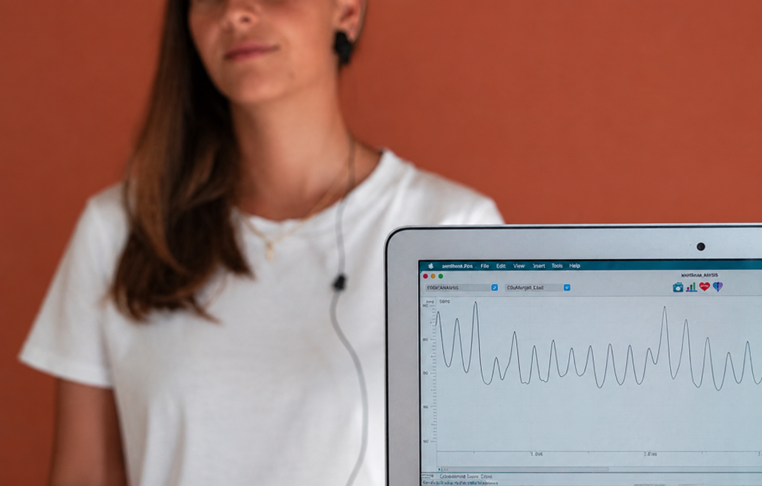 Vrouw met lang bruin haar draagt een witte T-shirt en kijkt naar een computerscherm met grafieken en analyse, tegen een oranje achtergrond.