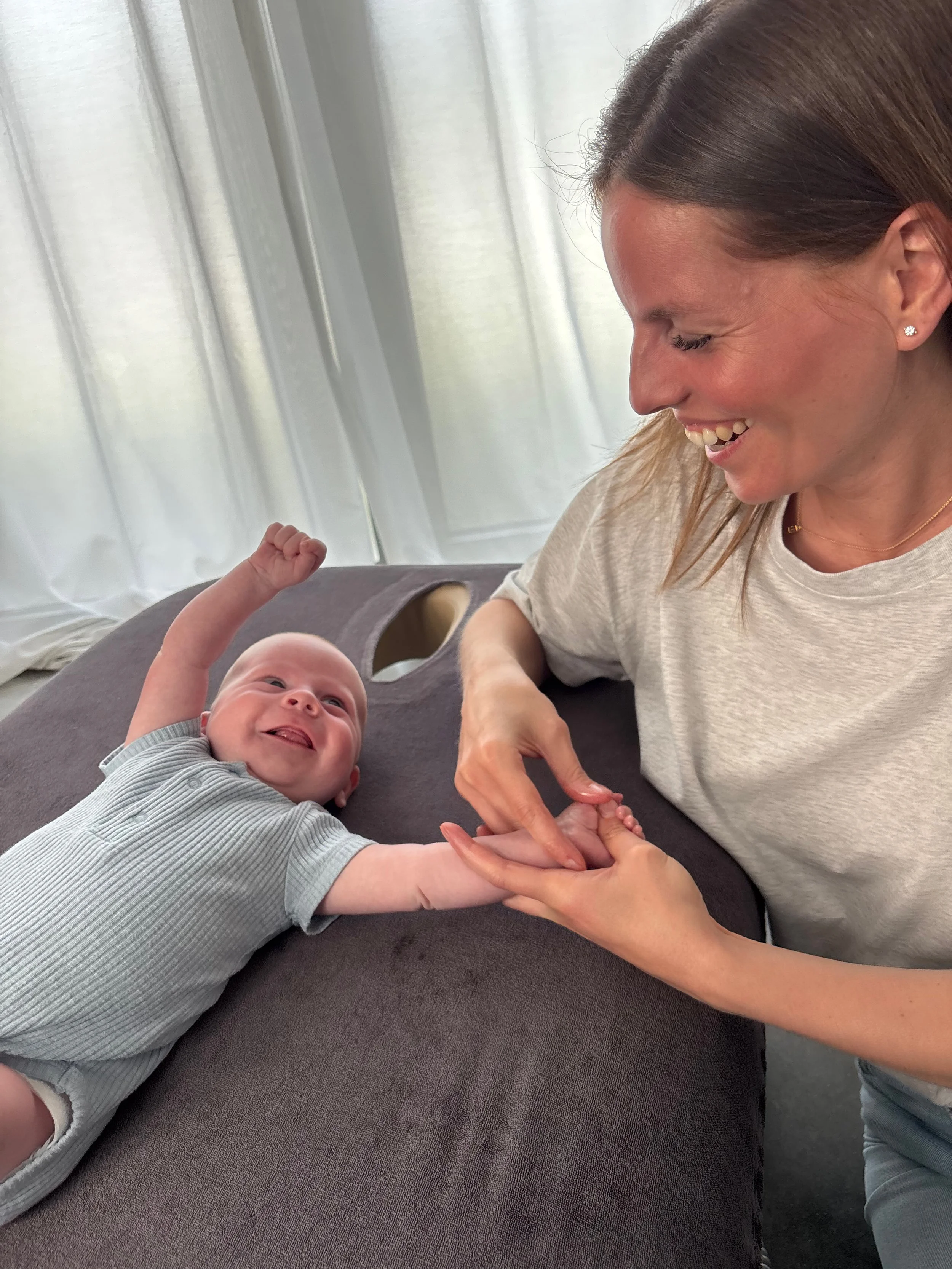 Een lachende vrouw en een baby die hand in hand liggen op een bankje, lachend naar elkaar.