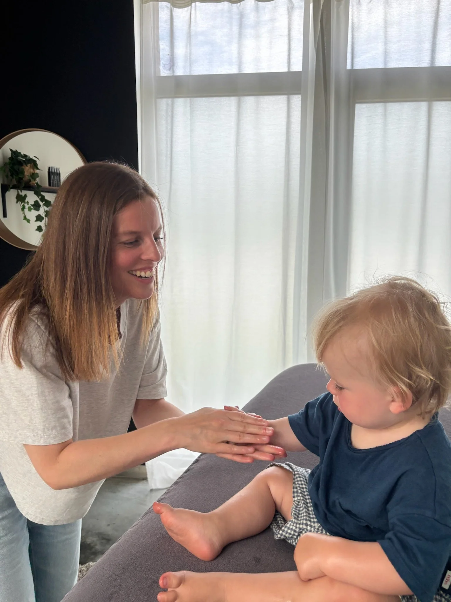 Een vrouw helpt een jonge jongen met een handvriendelijke activiteit op een tafel in een kamer met witte gordijnen en een muur met een ronde spiegel en planten.