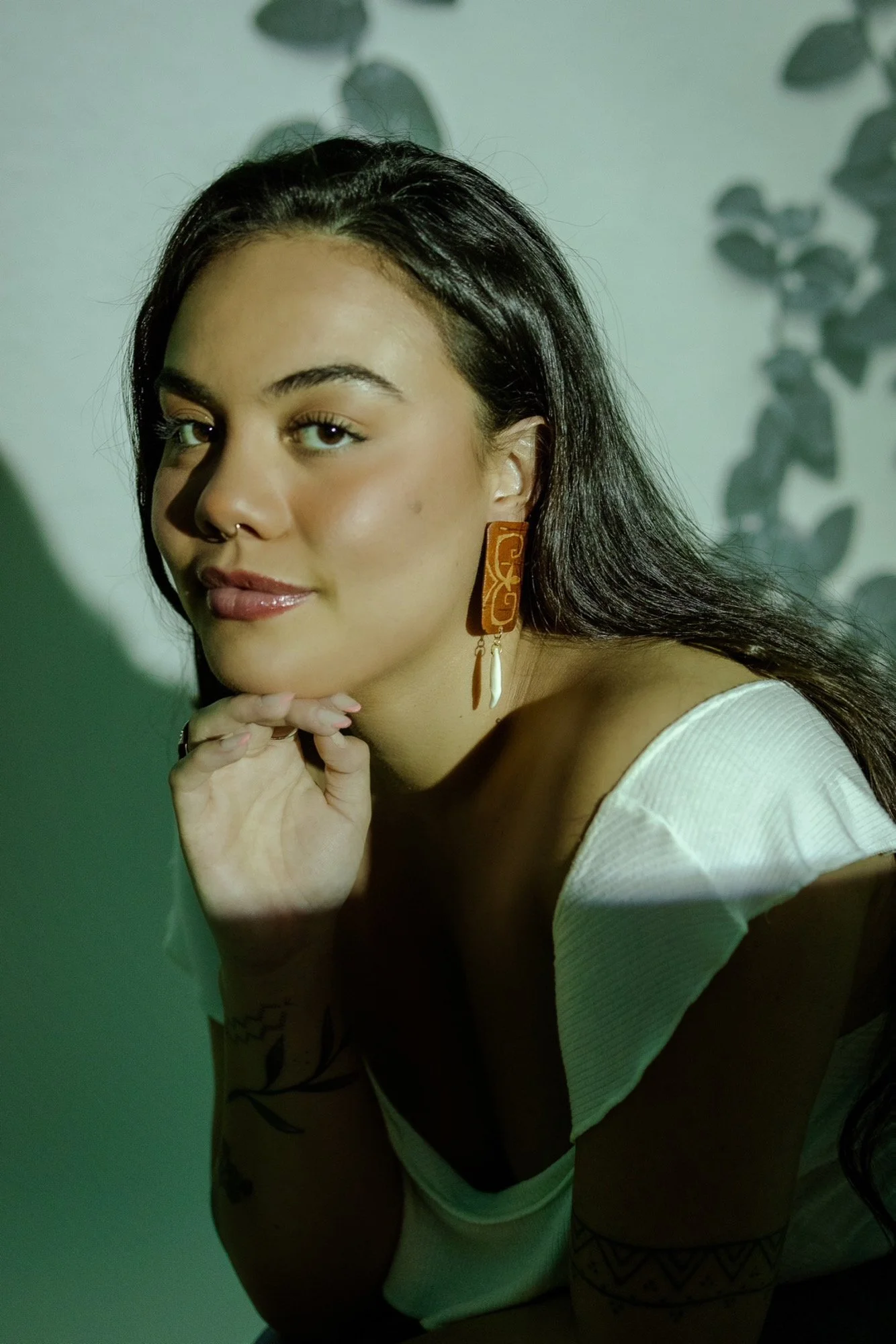 Kaitlin young woman with dark hair, wearing large earrings, resting her chin on her hand, looking at the camera. She has tan skin, a septum piercing, and tattoos on her left arm. She is wearing a white top, and the background has shadows of leaves.