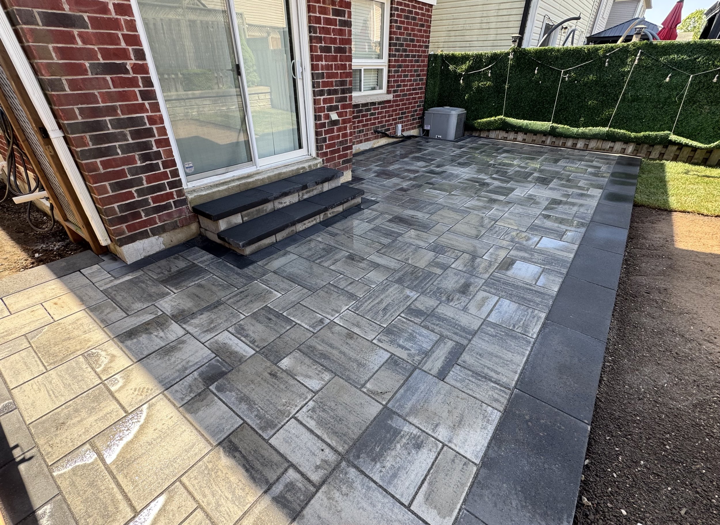 A backyard patio with gray and beige brick-patterned pavers, a sliding glass door, and a green privacy hedge. There is a small step with black tiles leading to the door, and a gray air conditioning unit is visible near the corner of the house.