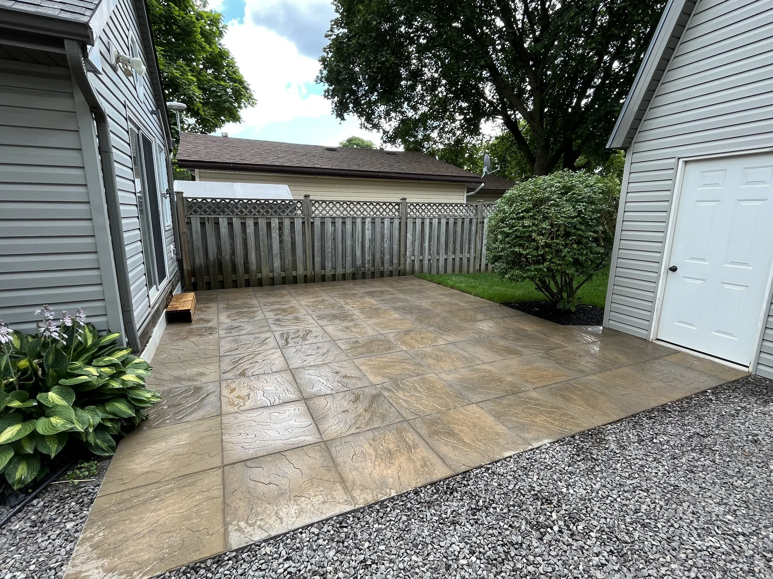 Backyard patio with wet stone tiles, a wooden fence, green bushes, a large tree, and neighboring houses with satellite dishes on the roofs.