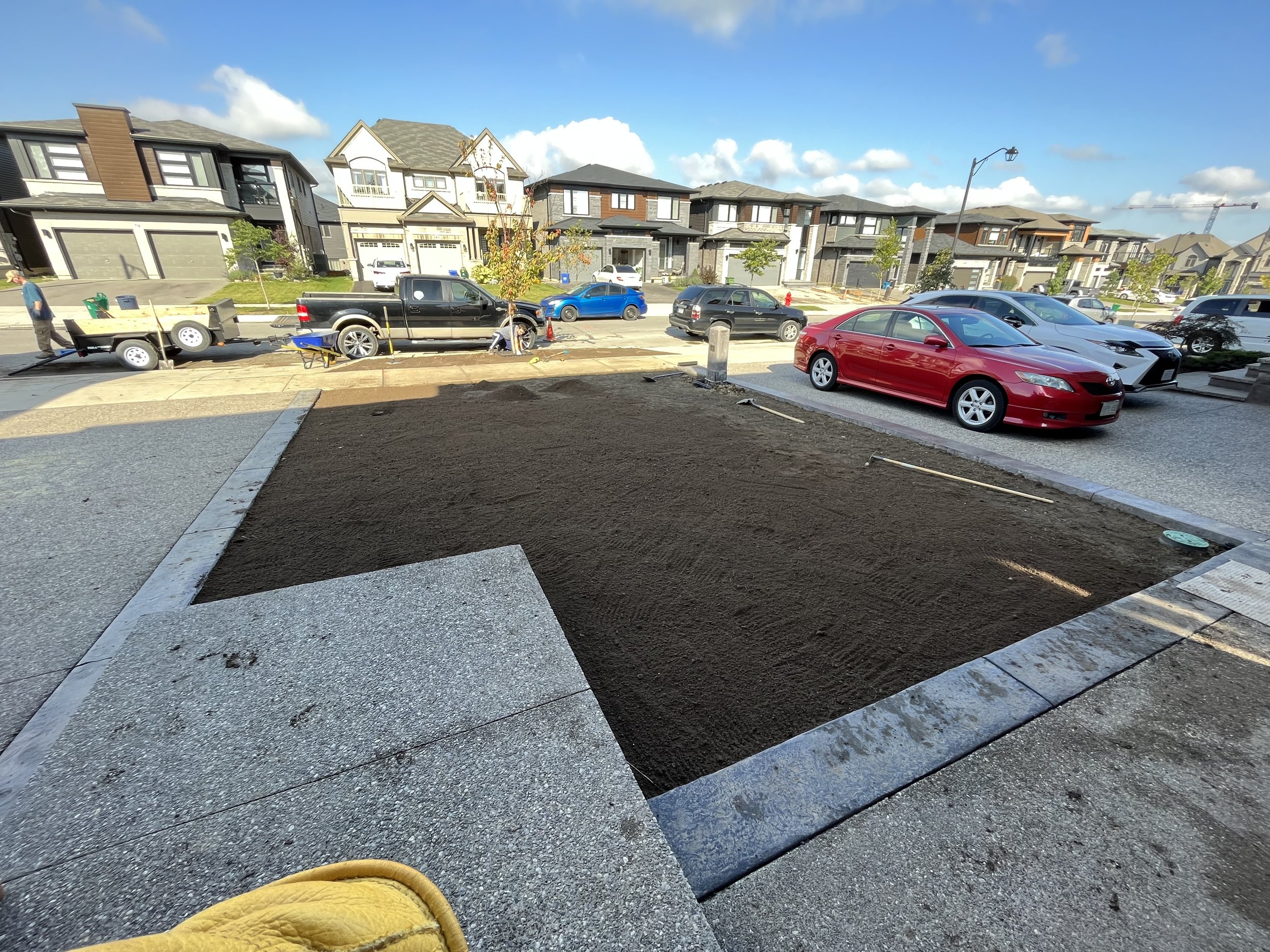 View from a construction site showing new sidewalk and driveway in a residential neighborhood with multiple modern houses and parked cars. A worker is visible near a trailer on the left side of the image.