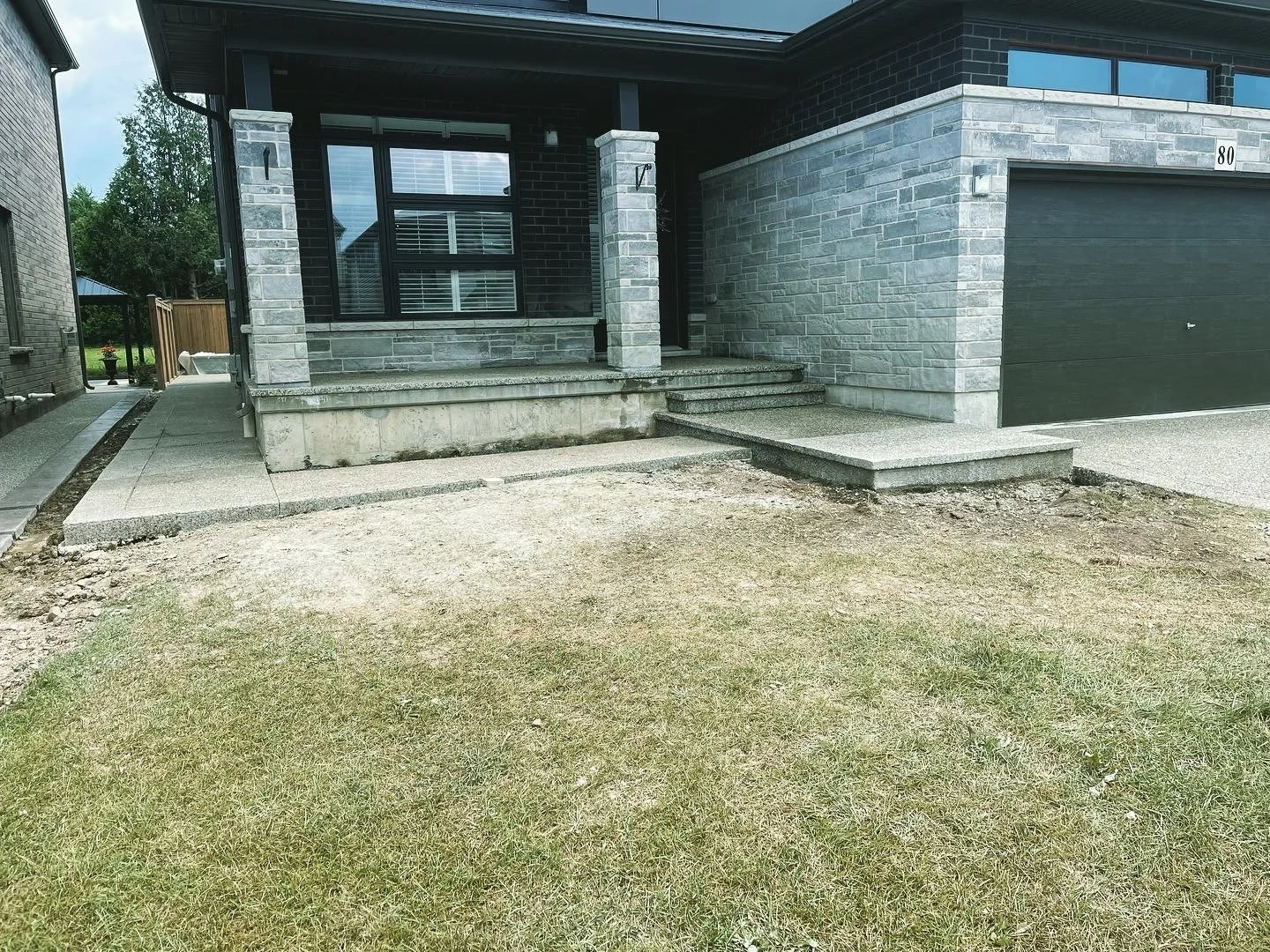 Front entrance of a modern house under construction with newly poured concrete steps and sidewalk, gray brick exterior, black garage door, and a small lawn area.