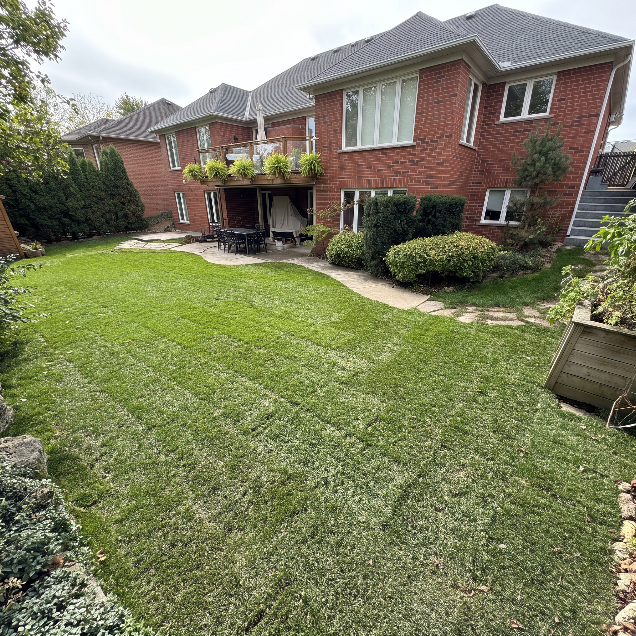 Backyard with green lawn, shrubs, trees, patio table with chairs, and a two-story brick house with large windows and a balcony.