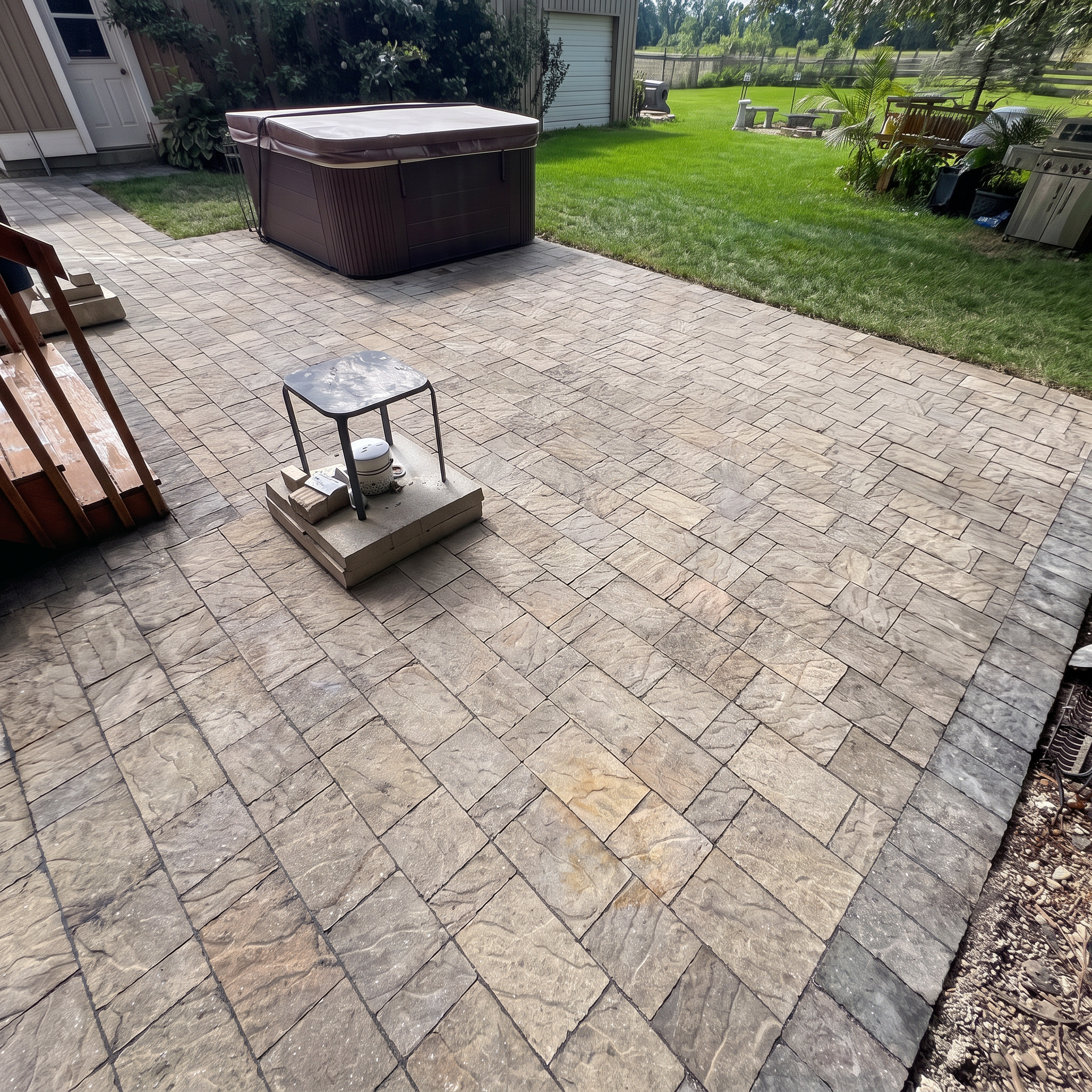 Patio area with beige paver stones, a hot tub with a brown cover, and outdoor furniture including a small table and bench, with a lawn, trees, and a fence in the background.