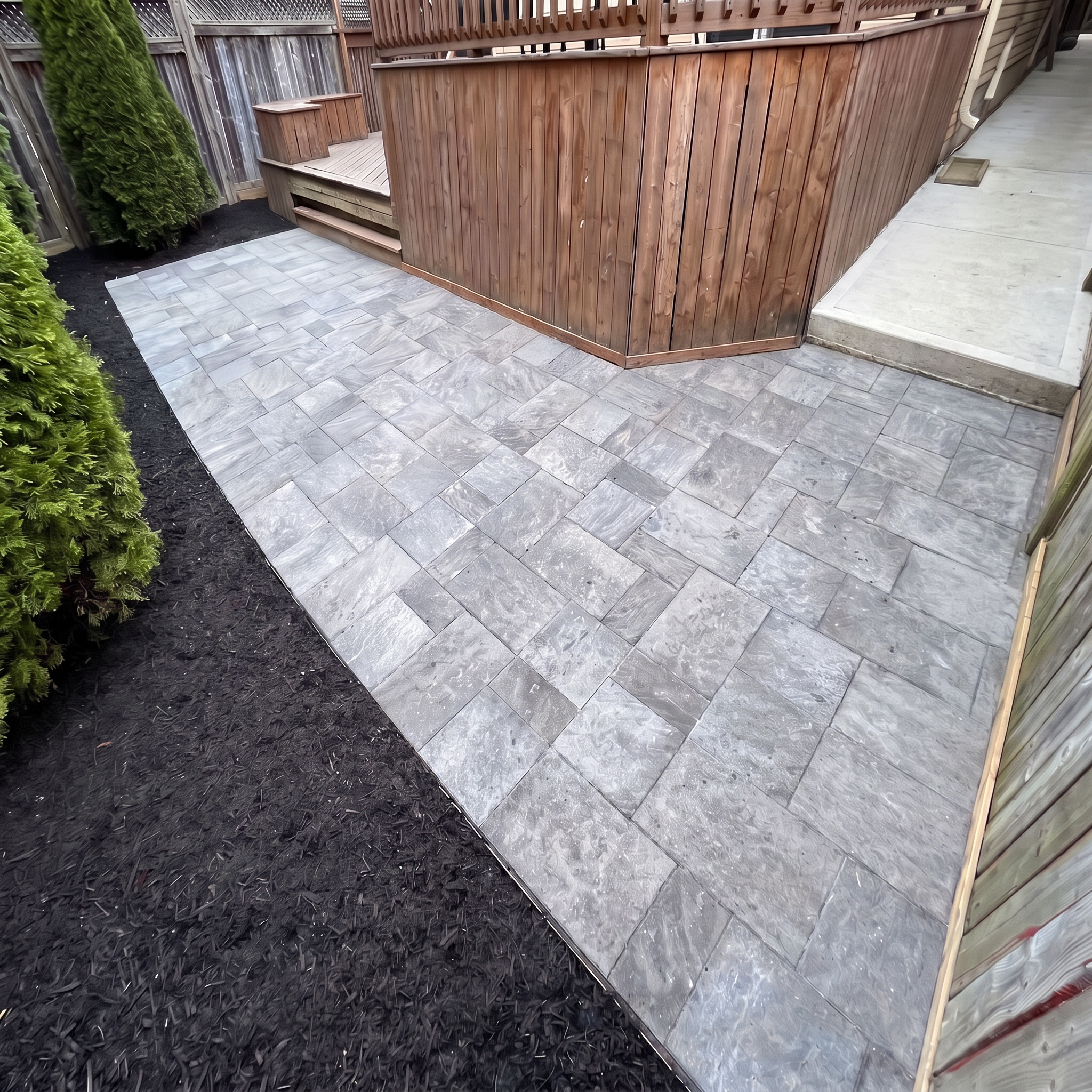 A backyard patio with light gray tiled flooring, a wooden privacy fence, and green bushes on the left side. A small wooden deck or platform with steps is visible in the corner.