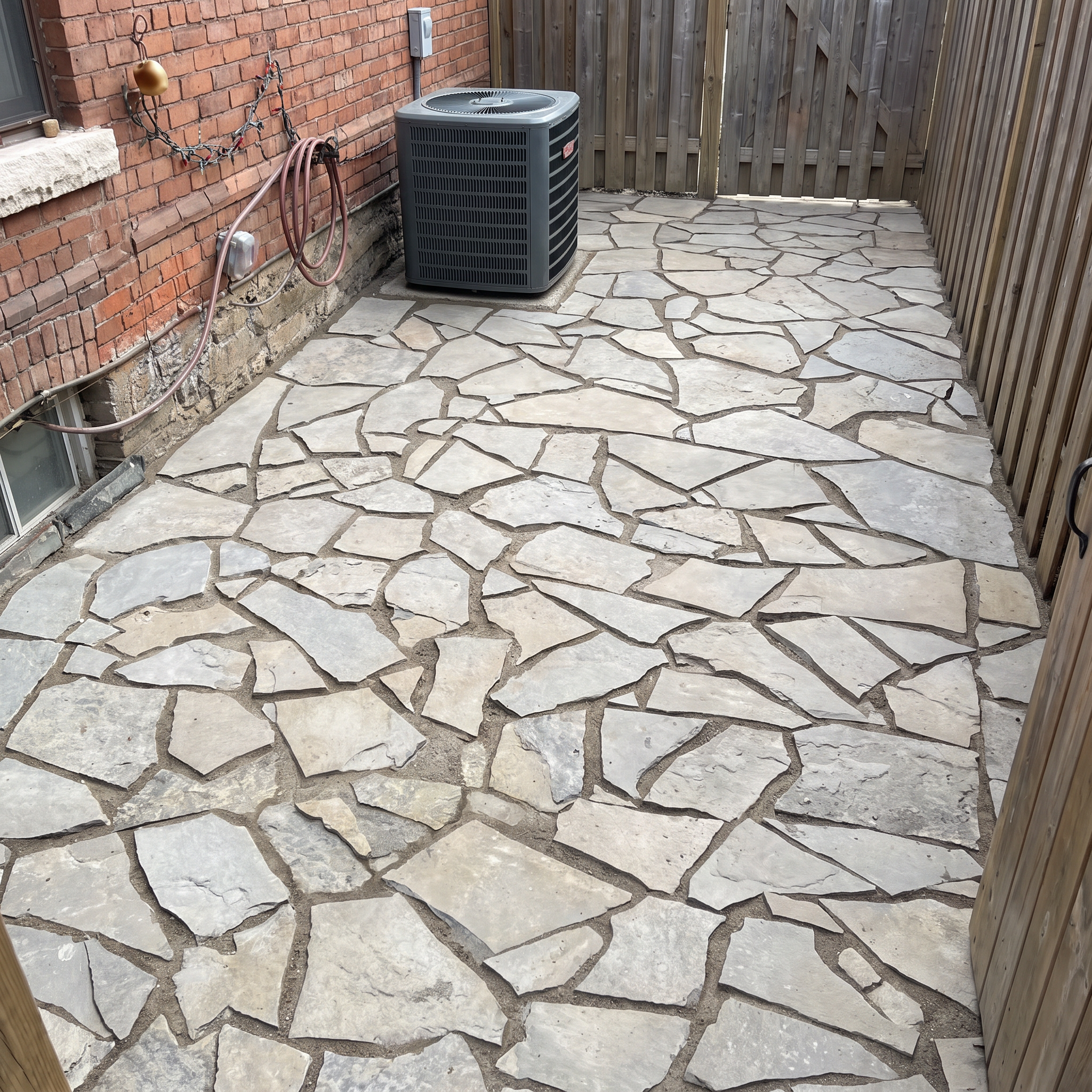 A small outdoor area with a stone tile floor, a brick wall on the left, a wooden fence on the right, and a gray air conditioning unit against the brick wall. There are some garden hoses and electrical connections nearby.