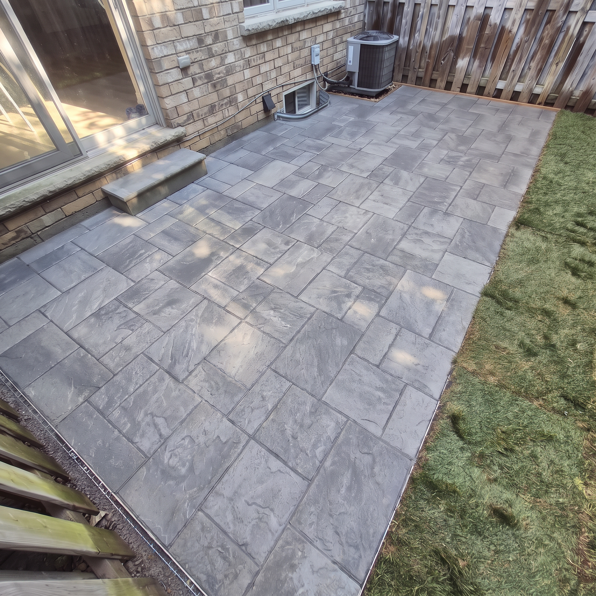 Backyard patio with stone tile flooring, brick house wall, sliding glass door, outdoor air conditioning unit, small window, wooden fence, and a patch of green grass.