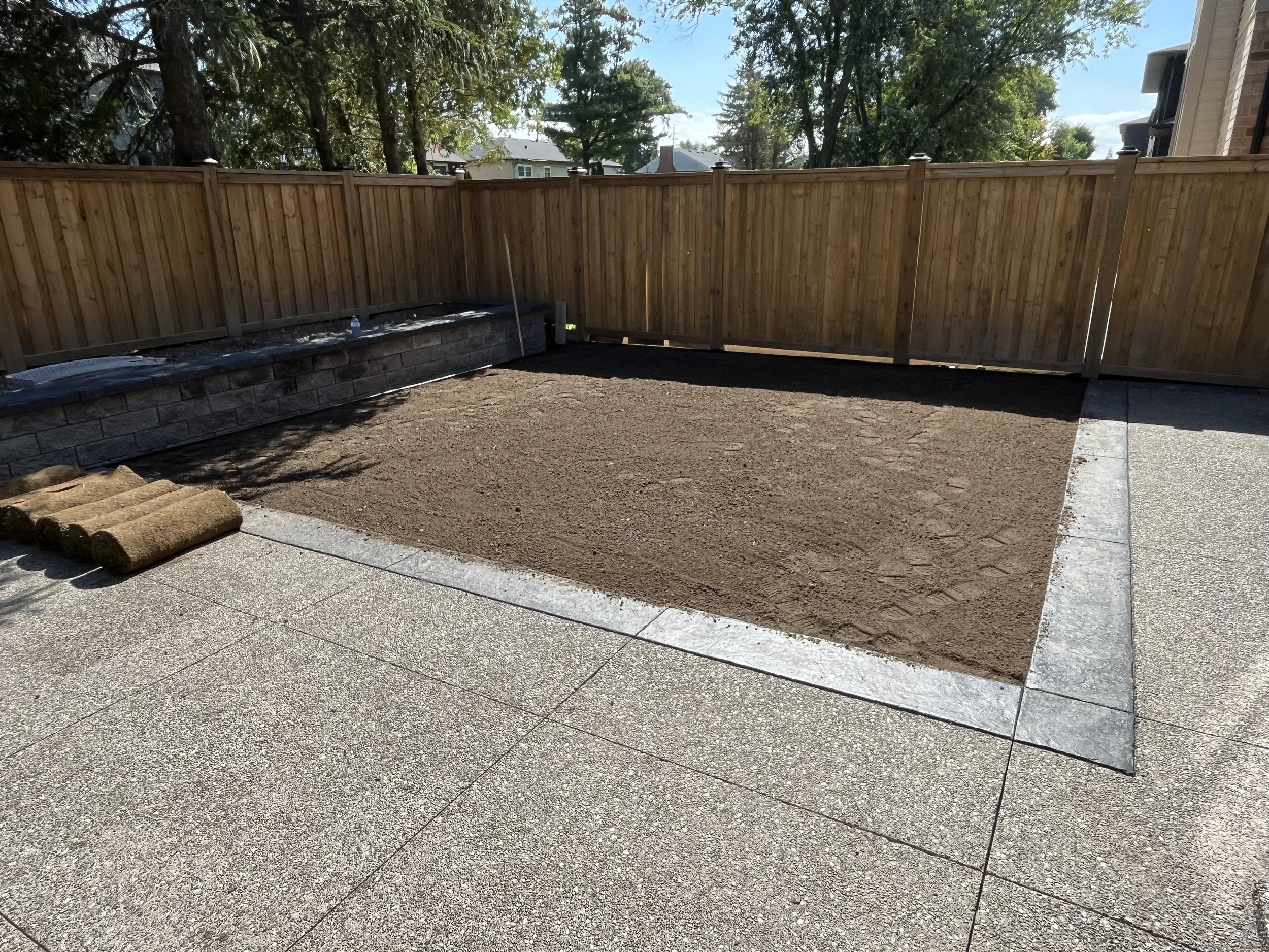 A backyard enclosed by a wooden fence. The ground appears to be prepared for landscaping or a garden bed, with some stones and a water bottle visible. Trees and neighboring houses are seen in the background under a partly cloudy blue sky.