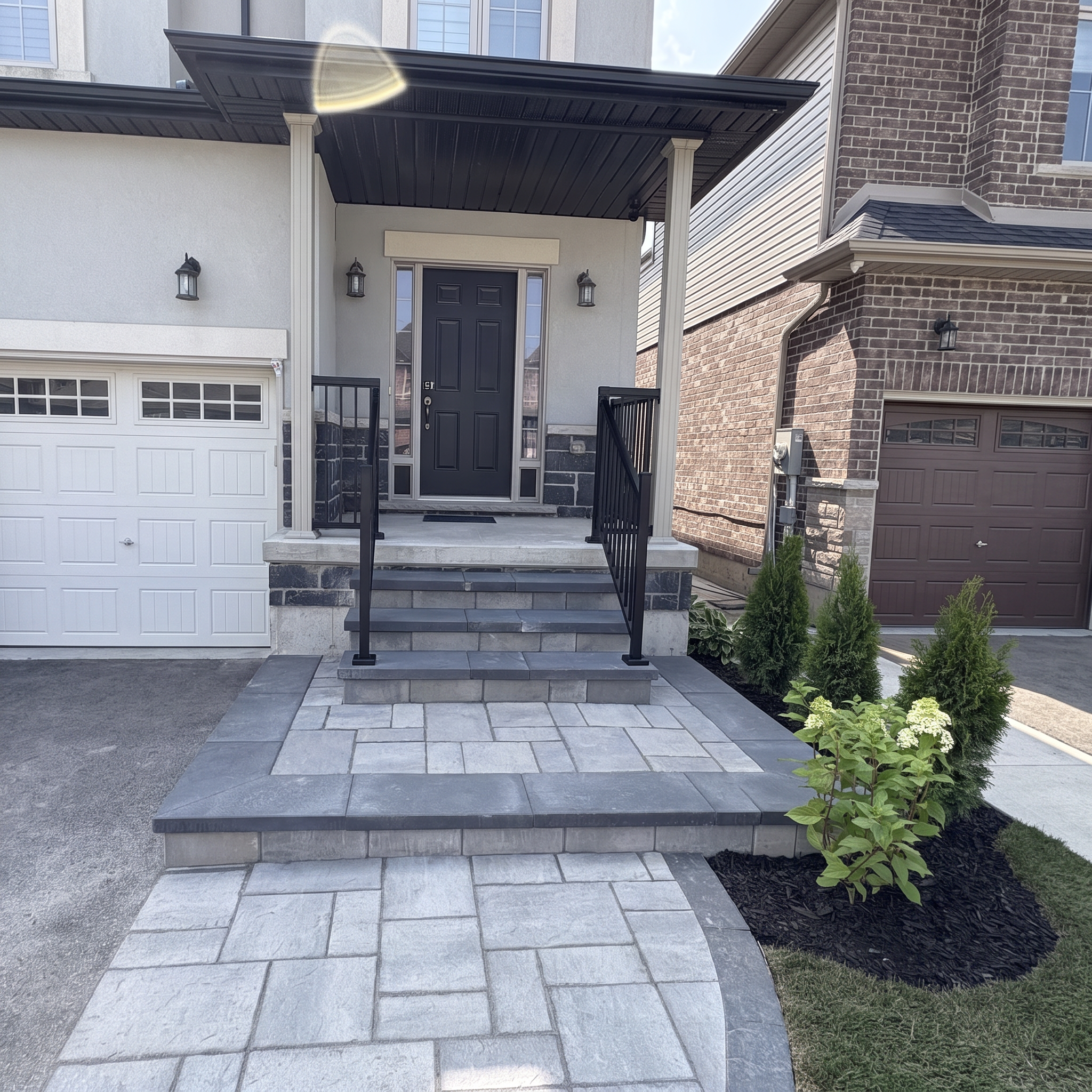 Front porch of a modern house with black door, black handrails, brick and siding exterior, two black lanterns, and a small garden with green plants.