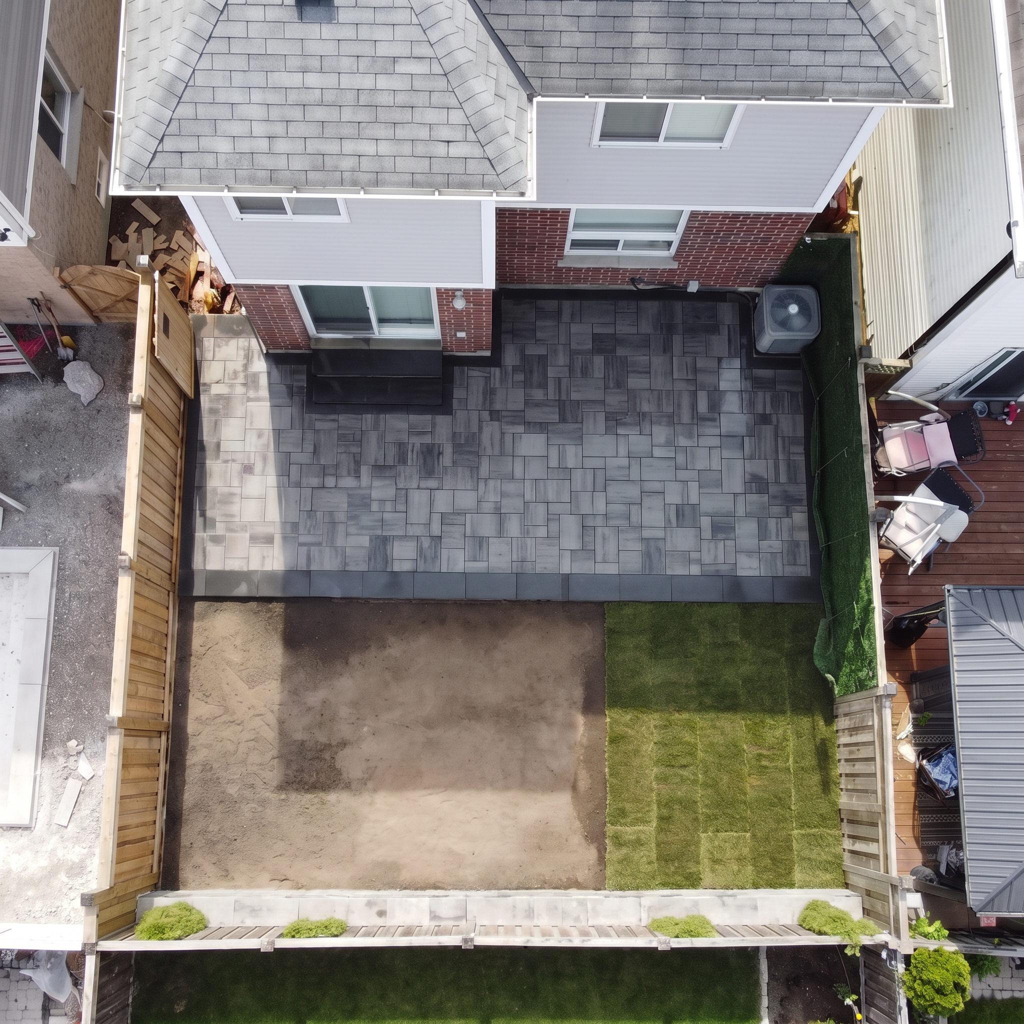 An aerial view of a backyard patio area with different types of flooring, including tile, concrete, and grass, surrounded by fences, with outdoor furniture and plants.
