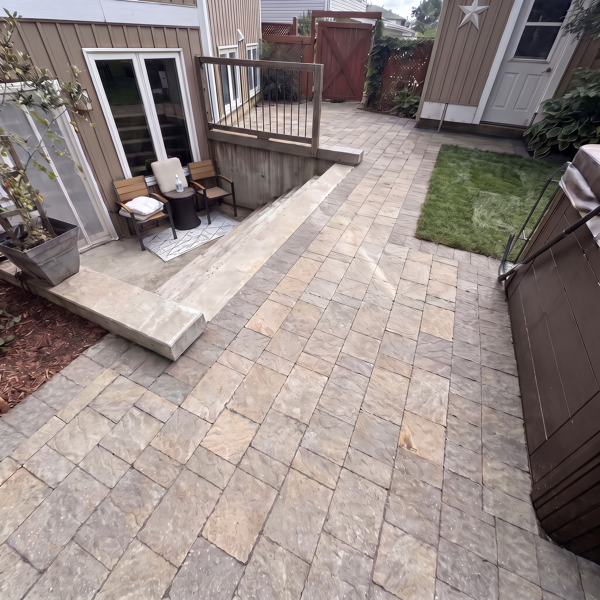 View of a backyard patio area with tiled flooring, a small seating area with chairs, a rug, and decorative stars on the wall, with a wooden gate and house exterior in the background.