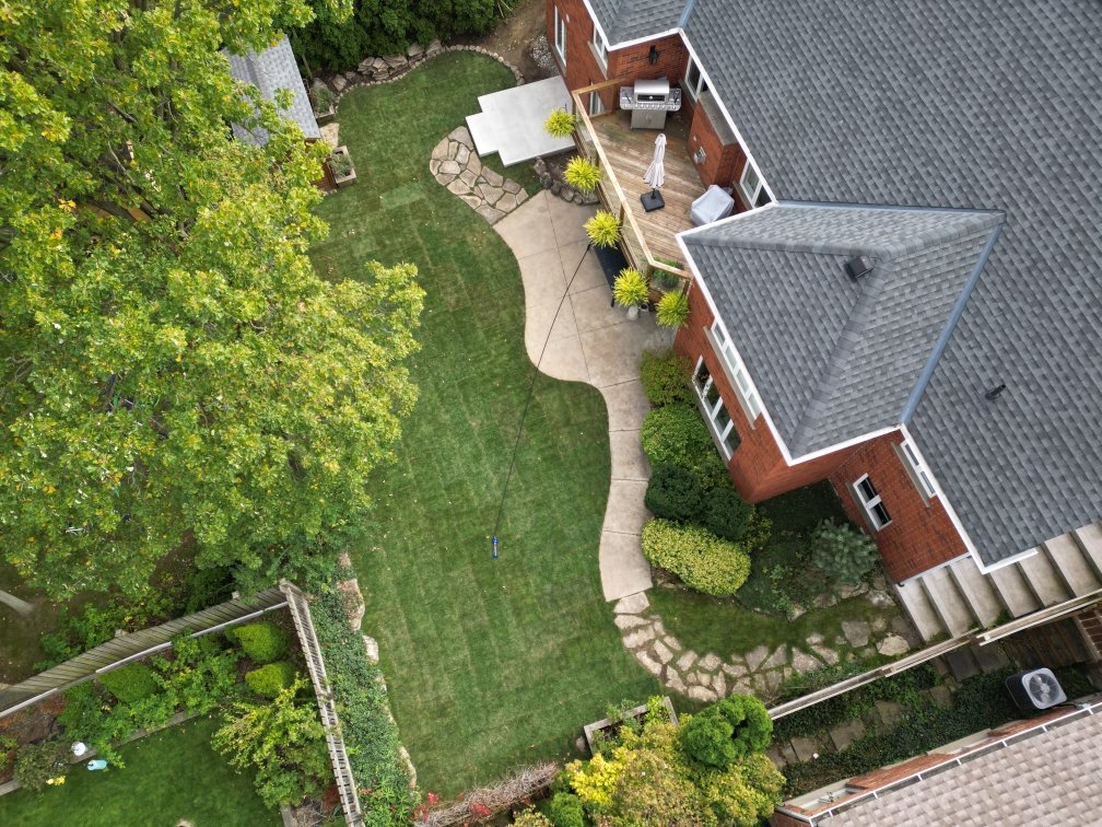 An aerial view of a backyard with a well-maintained lawn, concrete and stone pathways, and various trees and shrubs surrounding a brick house with a rooftop deck.