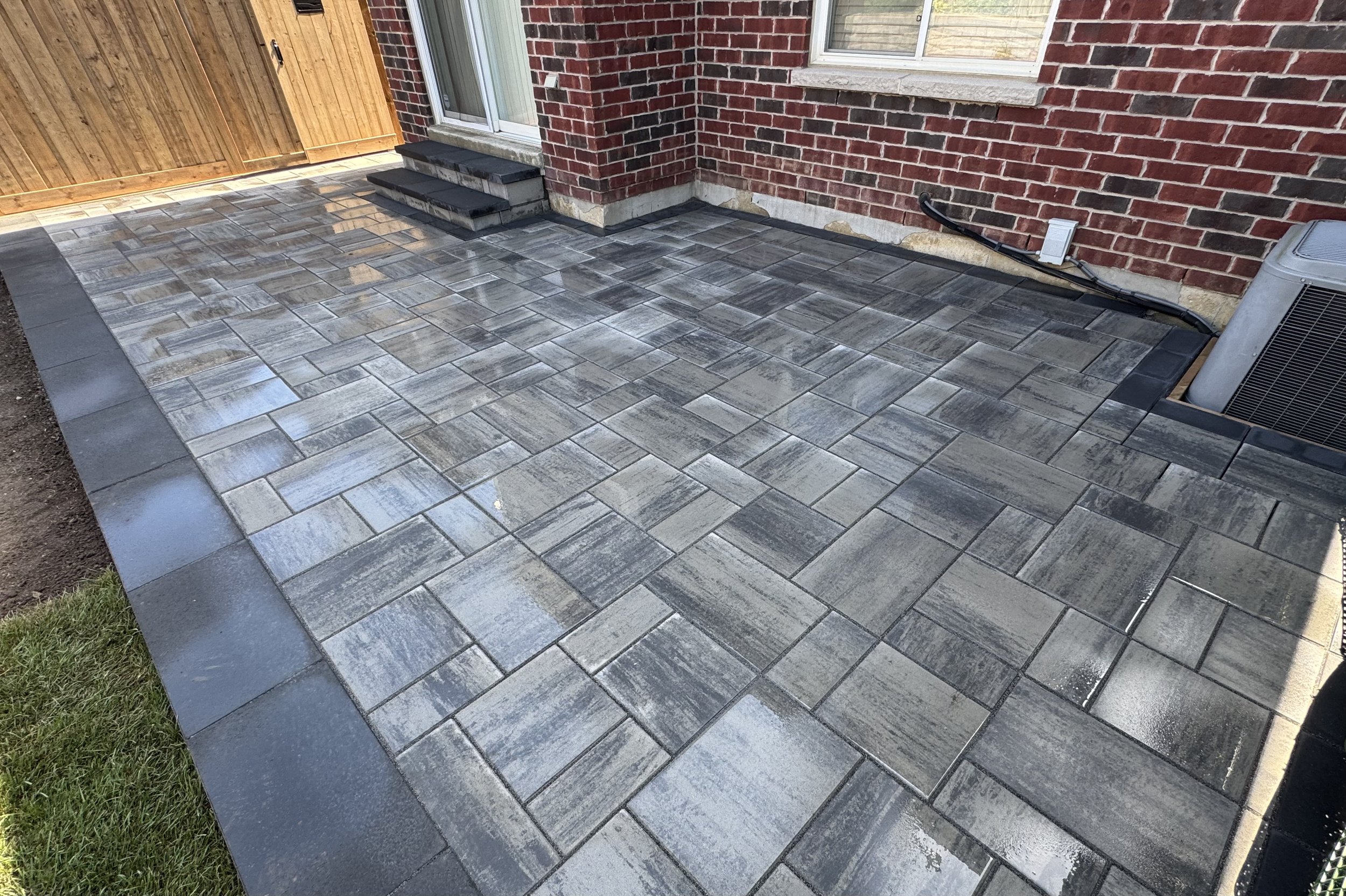 A backyard patio with gray stone tiles, a brick house wall, a sliding glass door, a small staircase, a wooden fence, an air conditioning unit, and electrical wiring along the wall.