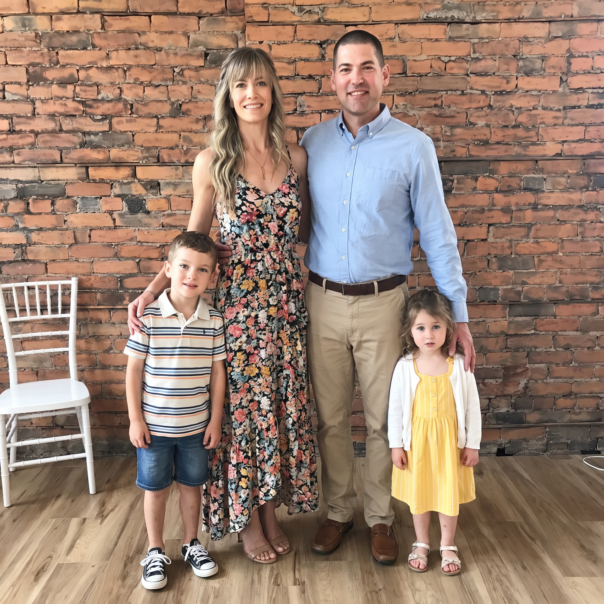 A family of five standing in front of a brick wall. The woman is wearing a floral dress, the man is in a light blue shirt and khakis, a boy is in a striped polo and shorts, and two young girls are dressed in yellow and white outfits.