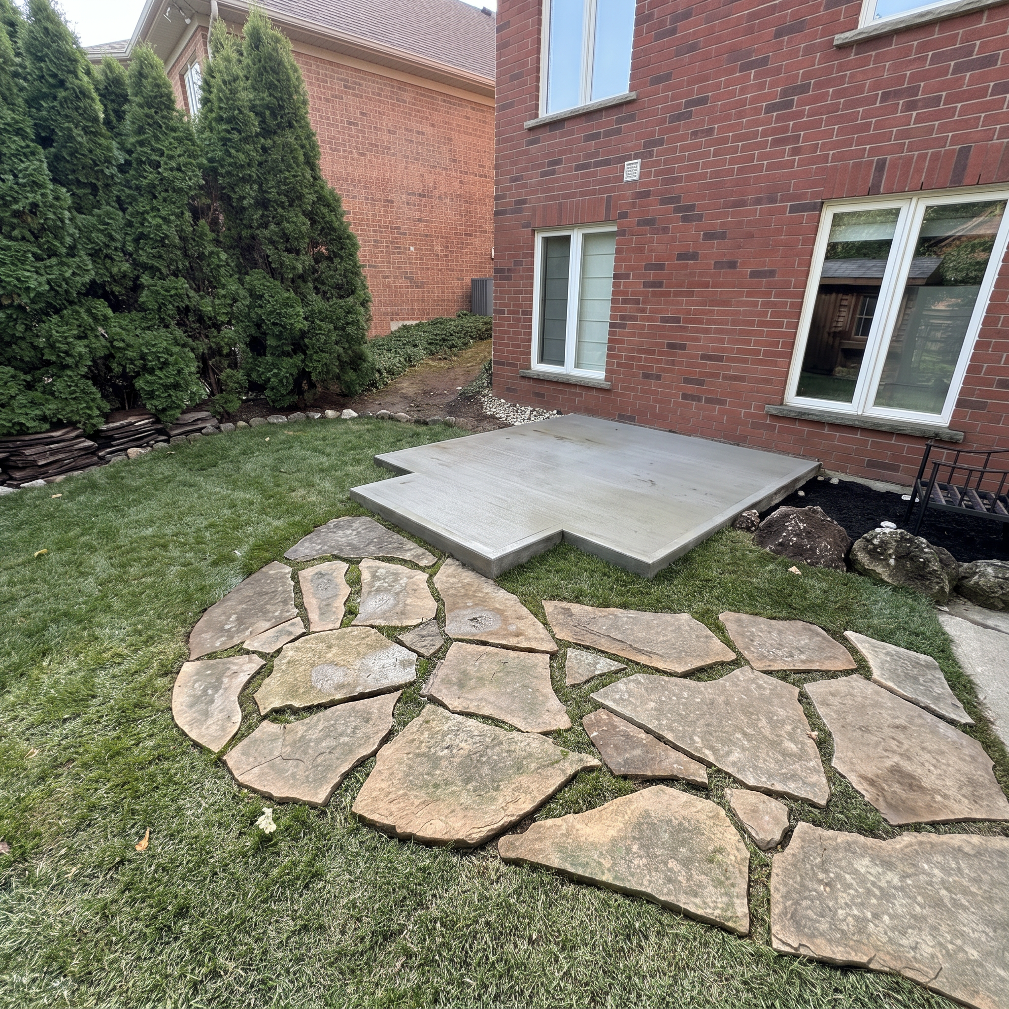 Backyard with brick house, large green bushes, stone pathway, small patio, and cement steps.