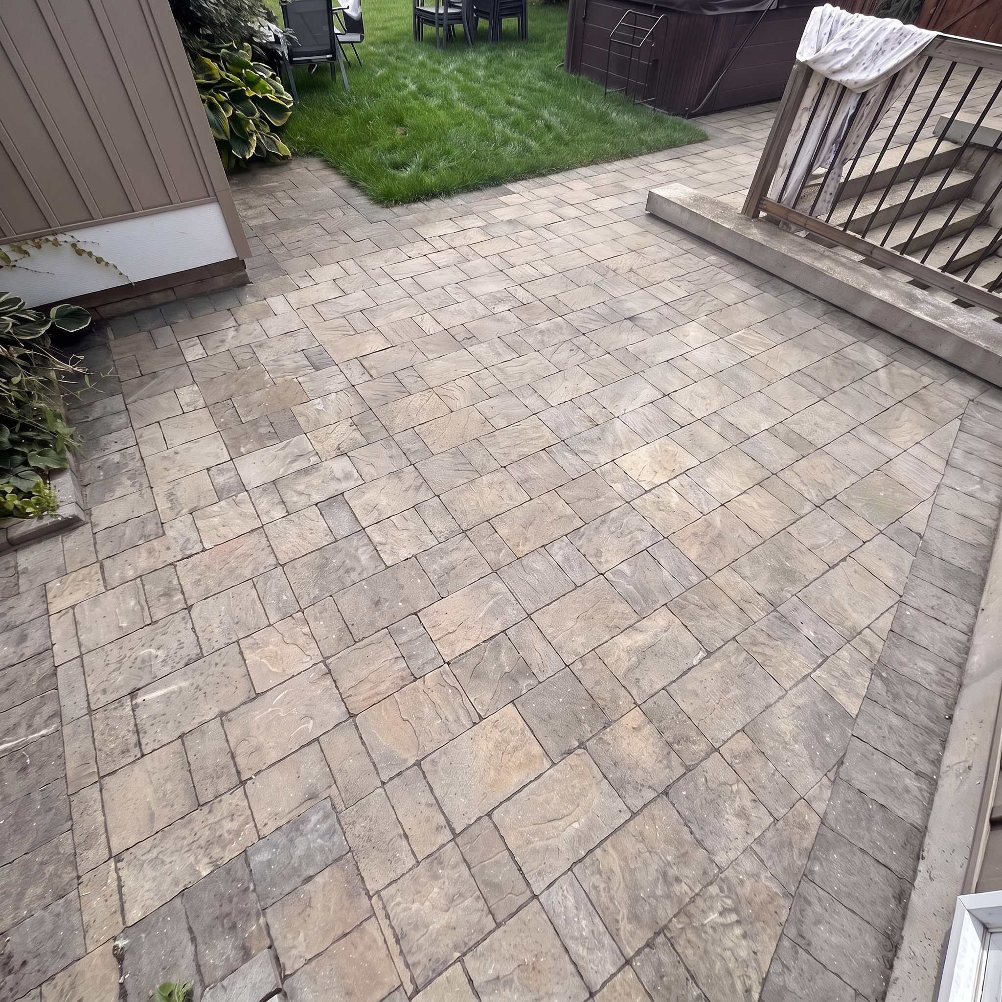 A backyard patio area with stone pavers, a small patch of grass, a metal railing staircase, outdoor furniture, and a shed or storage building in the background.