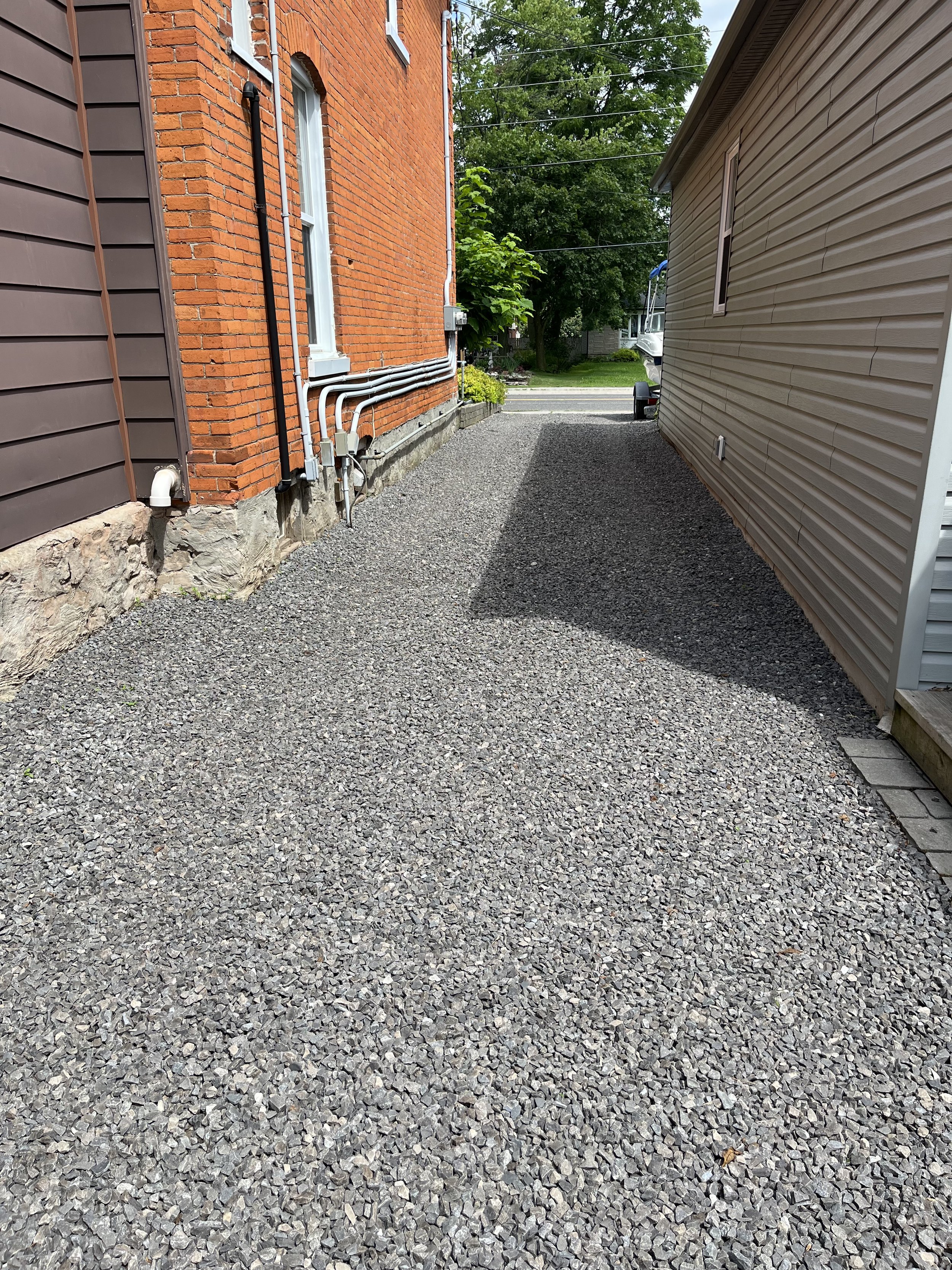 A gravel driveway between two houses, one brick and one vinyl siding, with a tree, some plants, and a small boat in the background.