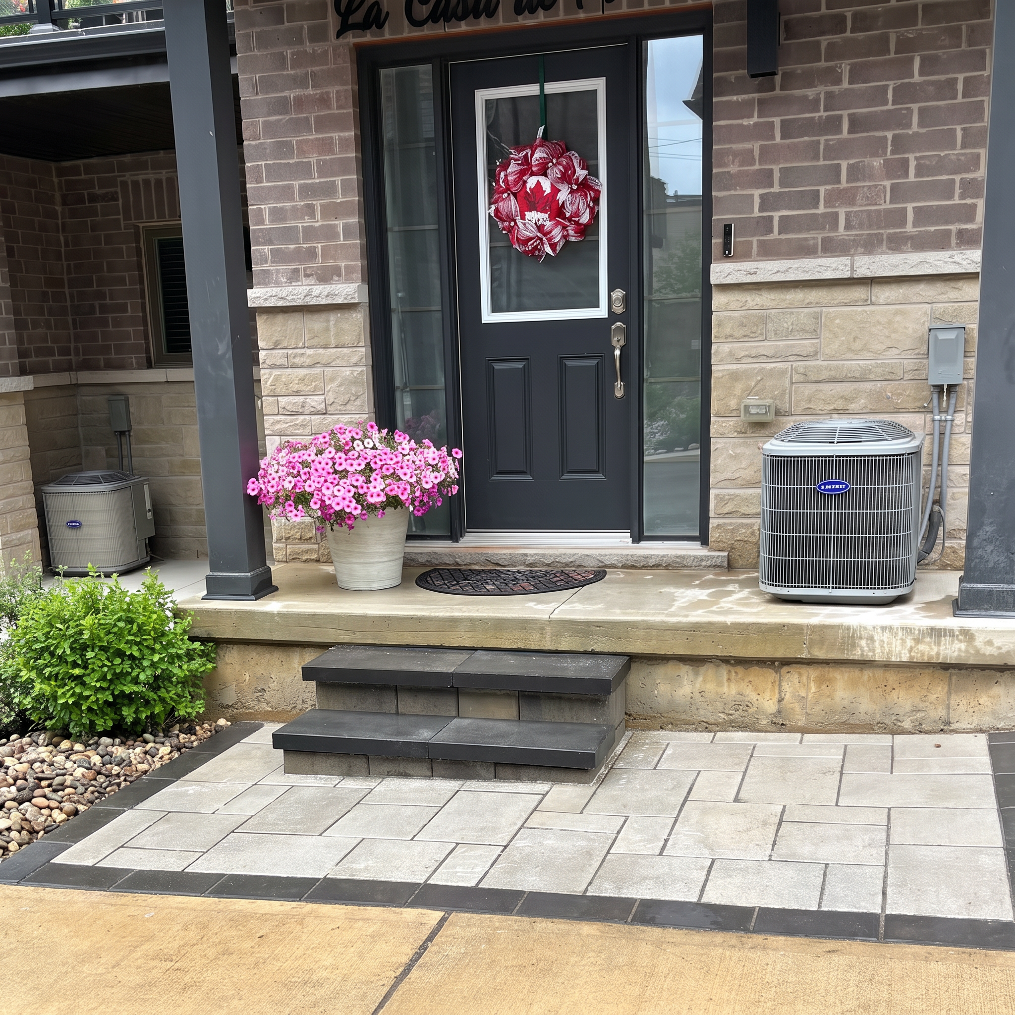 Front porch entrance of a house with a black door, a decorative red and white wreath, pink flowers in a large white pot, an air conditioning unit, and steps leading to the door, with brick and stone exterior walls.