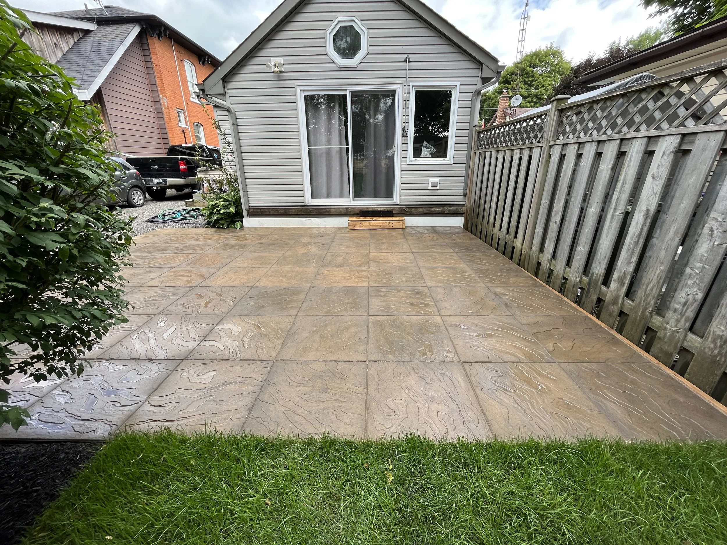 Backyard patio with tan stone tiles, bordered by green grass, and surrounded by a wooden fence on one side and plants on the other. A small wooden block is placed at the base of a light gray house with sliding glass door and window. Several cars are parked nearby, and a neighboring house with orange brick facade is visible in the background.
