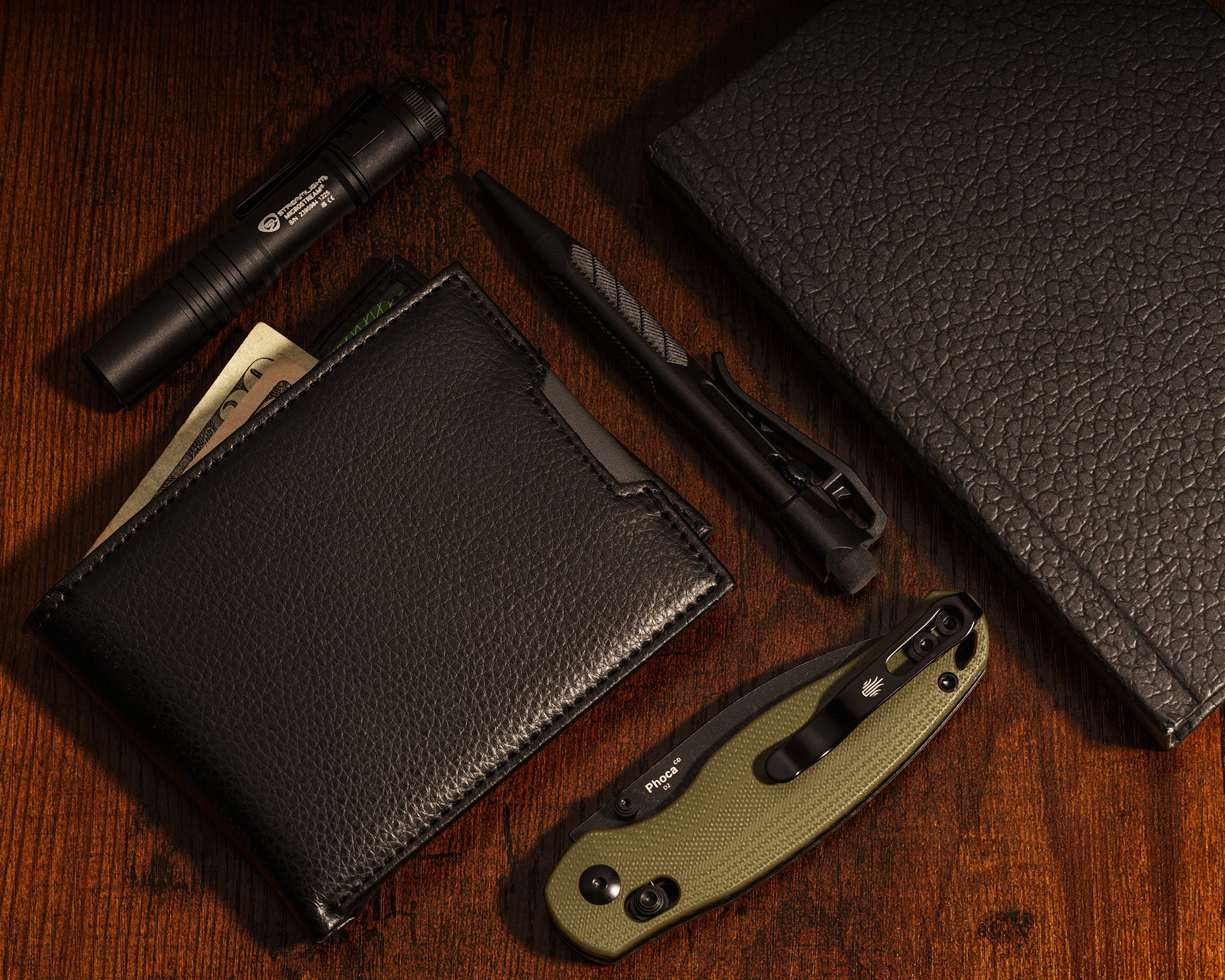 A tactical flashlight, a black leather wallet, a black pen, a black and silver multi-tool, and a tactical fixed blade knife with a green handle, all arranged on a dark wooden surface with a textured black box.