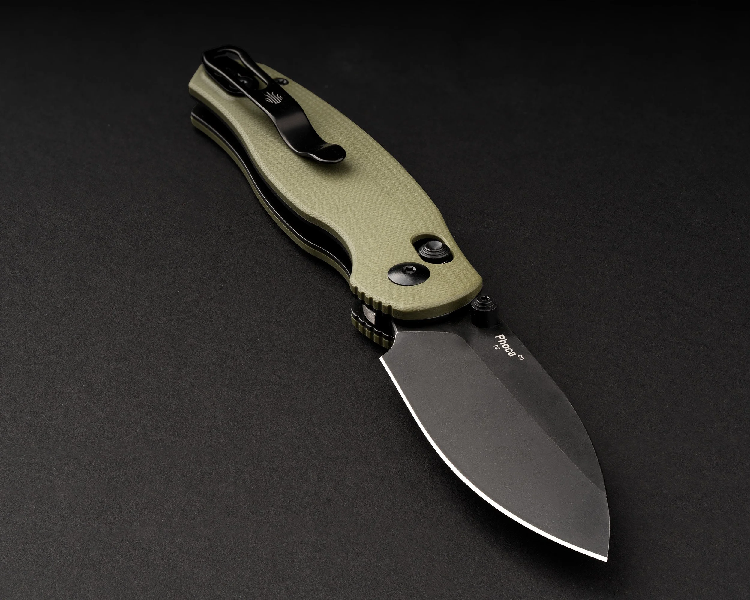 Close-up of a tactical folding knife with a black blade and olive green handle, resting on a dark surface.