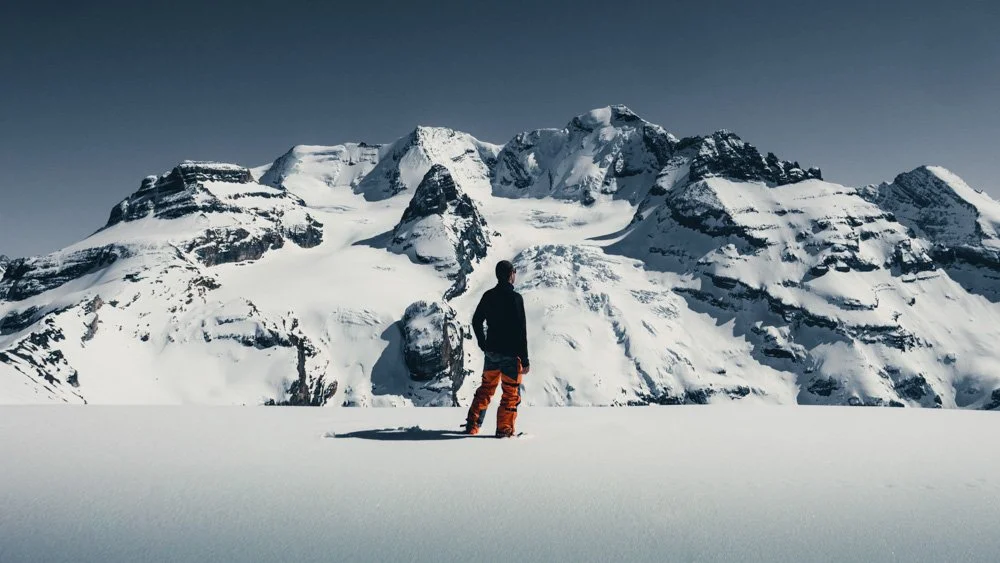 Ein einzelner Mensch steht auf schneebedecktem Boden vor einer grossen schneebedeckten Bergkette unter einem klaren Himmel.
