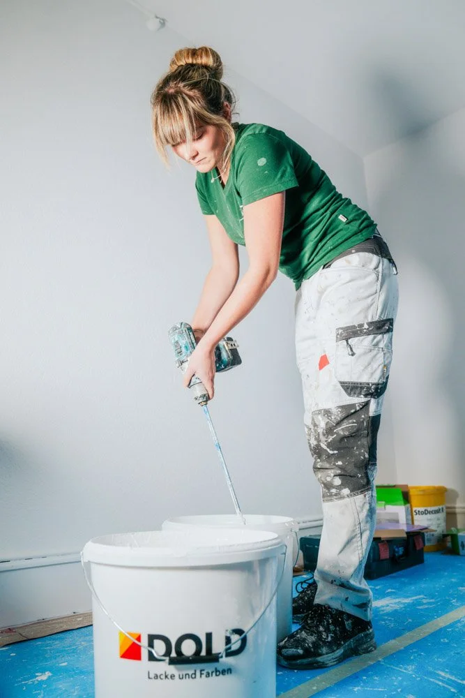 Frau beim Streichen einer weißen Wand, stehend vor einem Eimer mit Farbe, in einem Raum mit blauer Unterlage und Werkzeugkisten im Hintergrund.