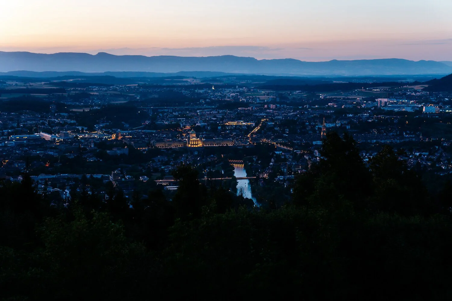 Blick auf eine beleuchtete Stadt während der Dämmerung, mit Fluss und Hügeln im Hintergrund.