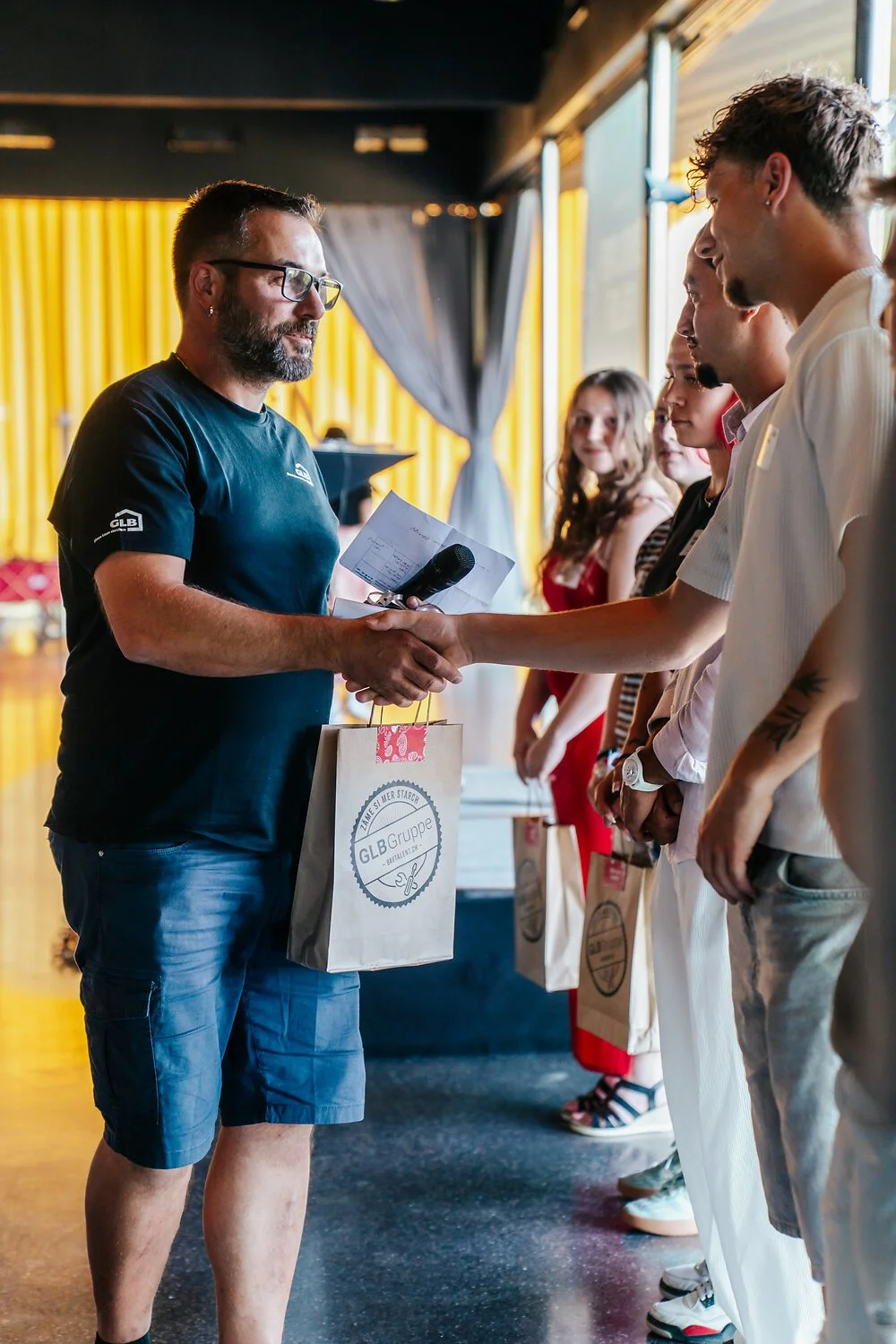 Ein Mann in schwarzem T-Shirt übergibt einem anderen Mann einen Preis bei einer Zeremonie, während eine Reihe von Menschen zuschaut.